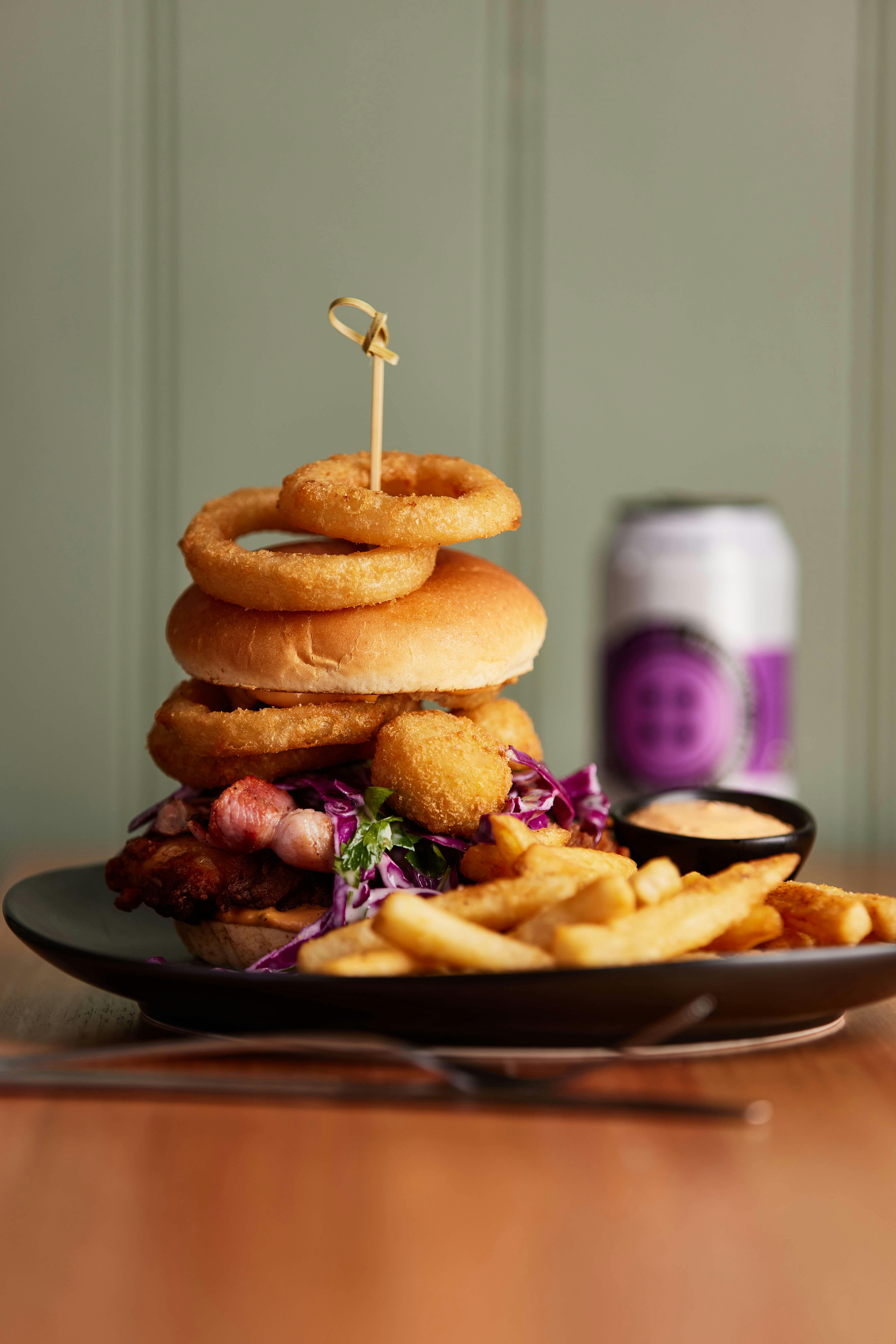 A tall burger topped with onion rings sits on a plate with chips and a craft beer in the background