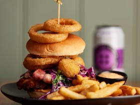 A tall burger topped with onion rings sits on a plate with chips and a craft beer in the background