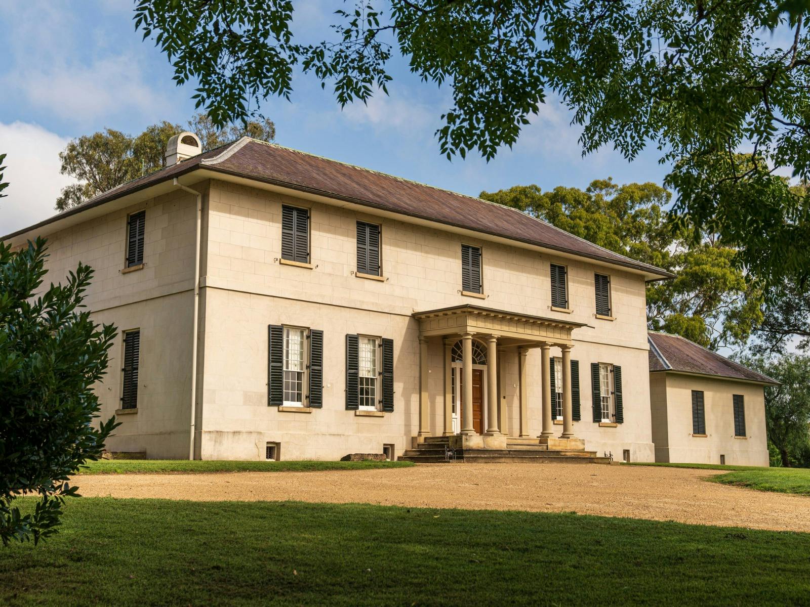 Double storey historic building faces the sunshine. Four pillars mark the front entry. Trees frame.