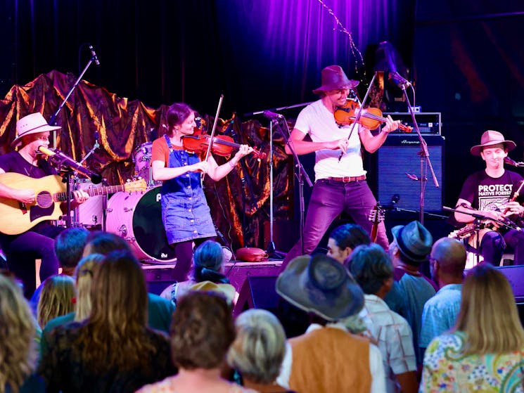 People performing onstage, two fiddler, guitar and Uillean pipes