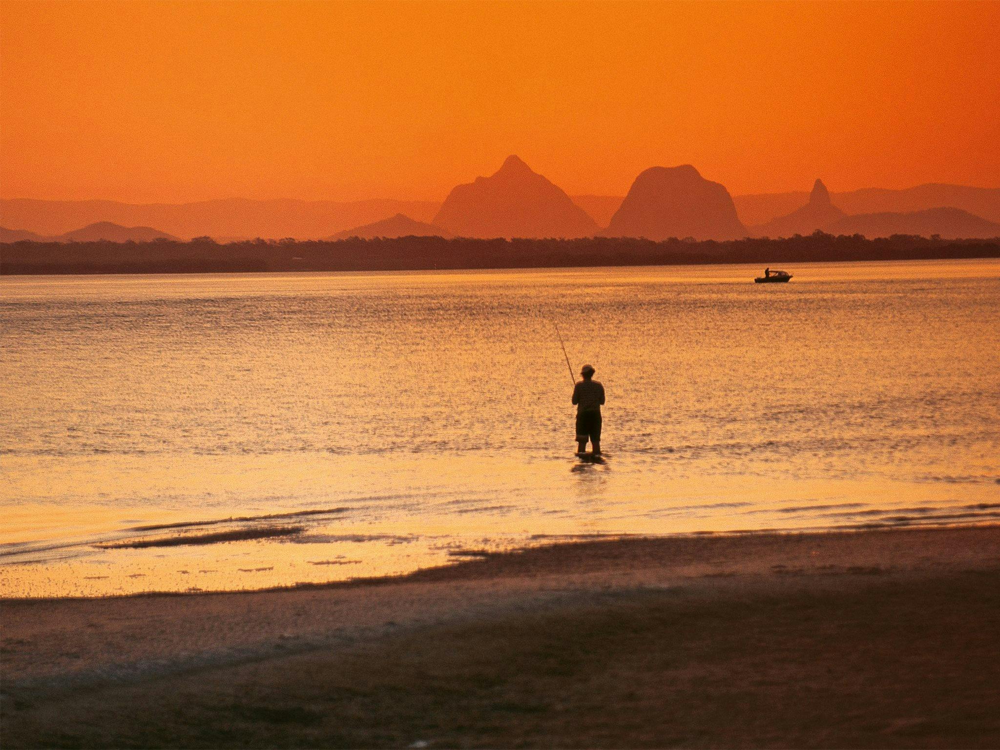 Oranged hued image of person standing in the ocean fishing with a boat further out and mountains Oranged hued image of person standing in the ocean fishing with a boat further out and mountains