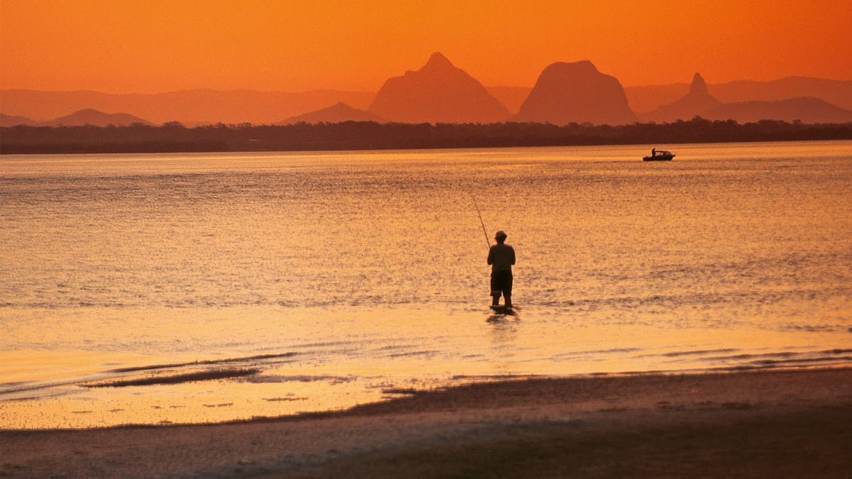 Oranged hued image of person standing in the ocean fishing with a boat further out and mountains