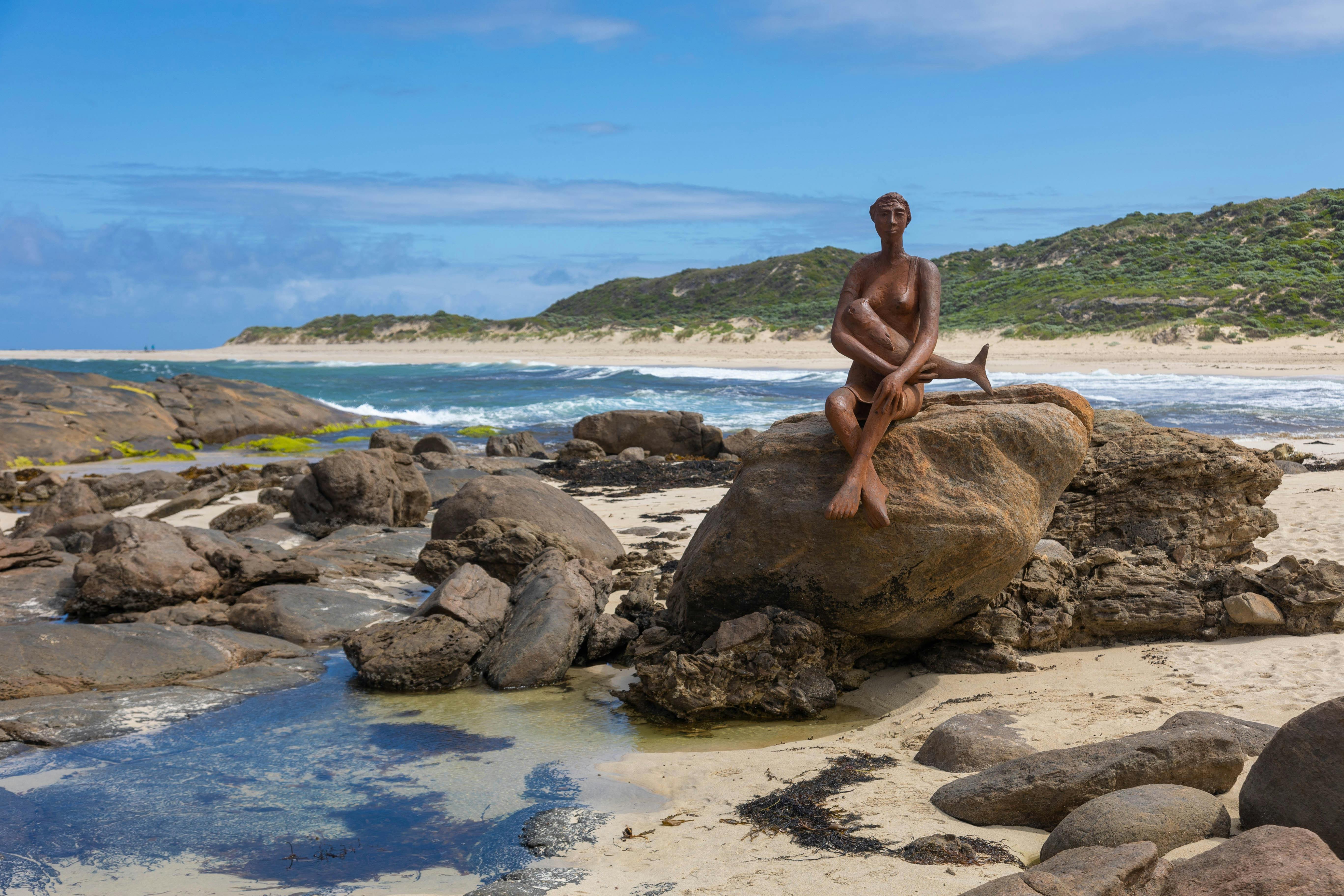 A stone mermaid on a rock with the coast in the background