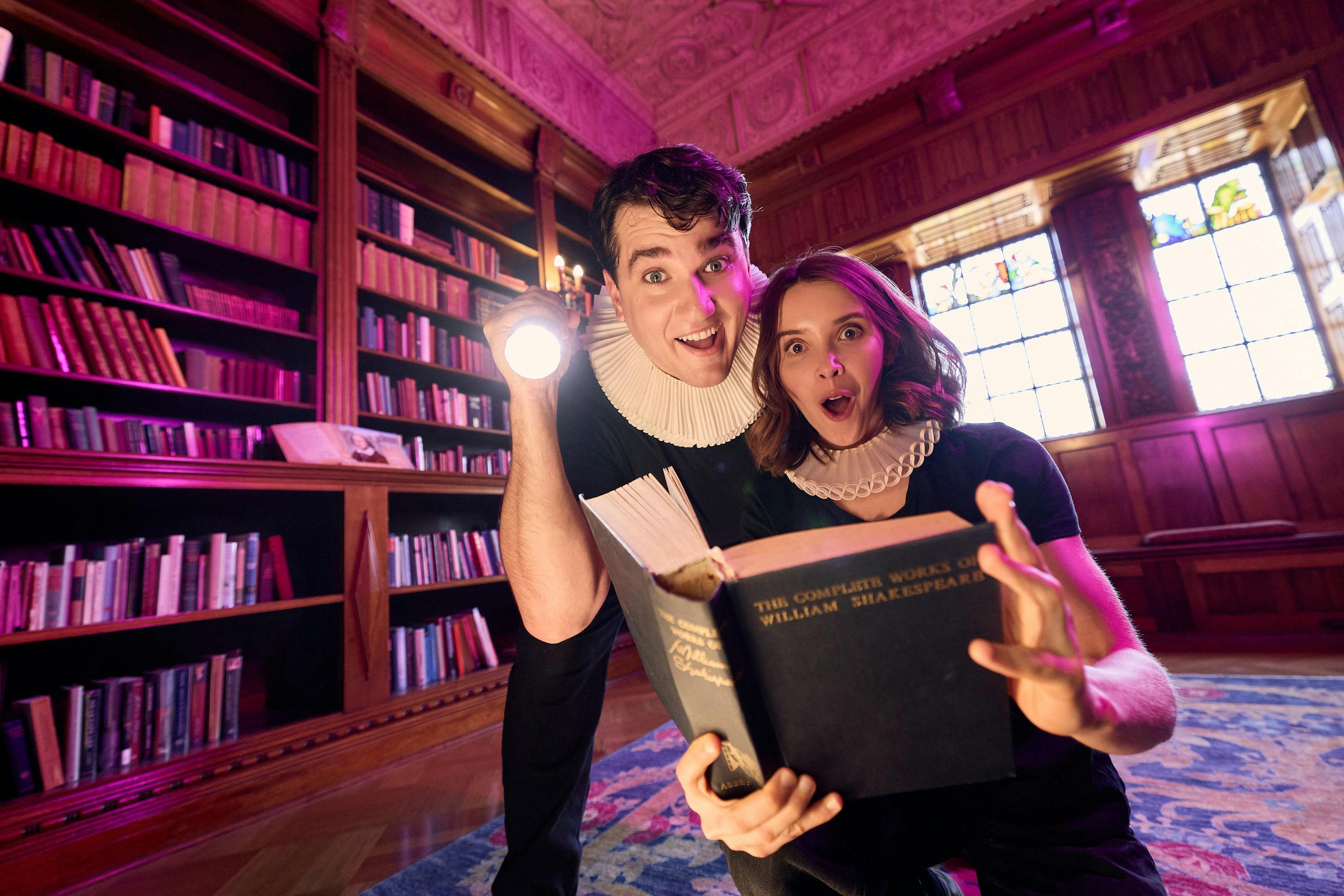 Two people in white neck ruffs holding a book and a torch and gesturing to the camera