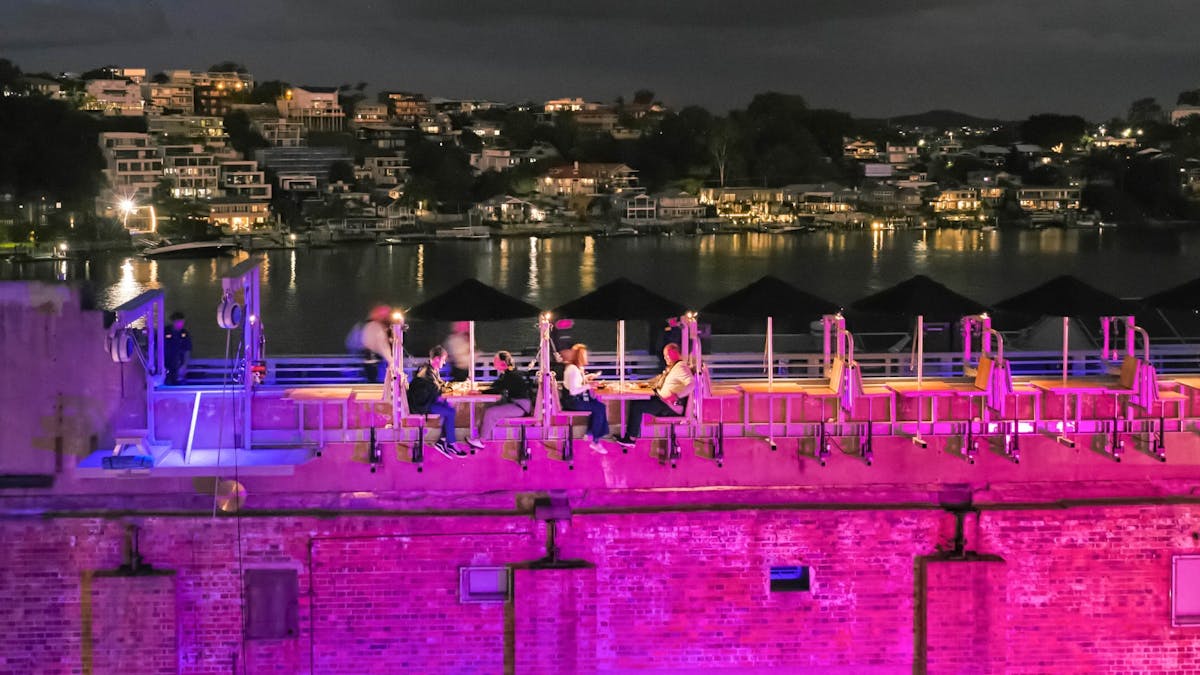 People dining on a rooftop above a lit pink brick building at night, overlooking a river.
