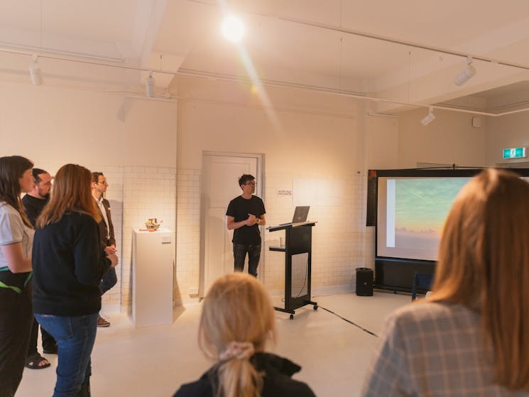 A man presenting in front of a projector in a gallery with an audience standing in a semi circle.