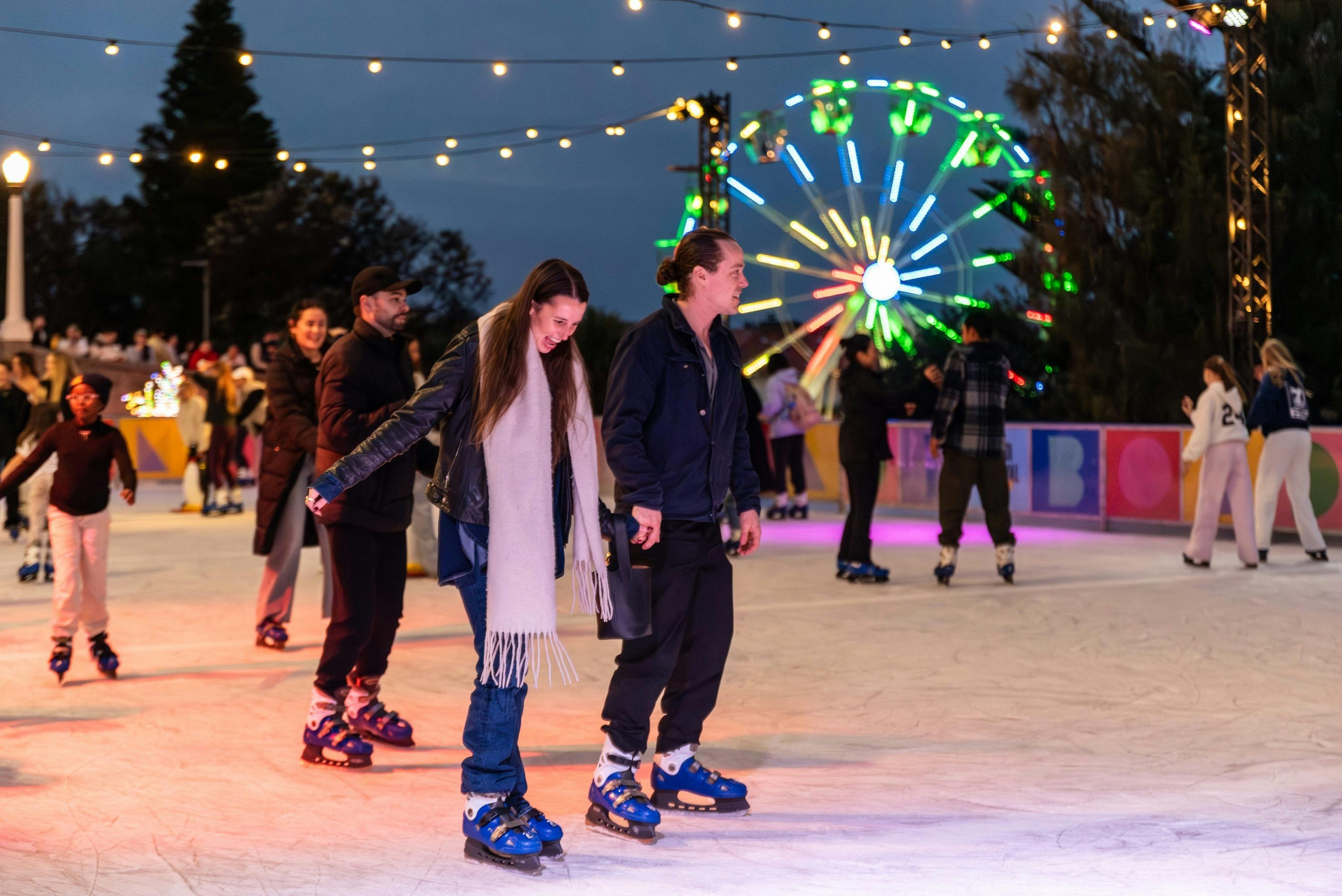 People ice skating under string lights at night, with a brightly lit Ferris wheel in the background.