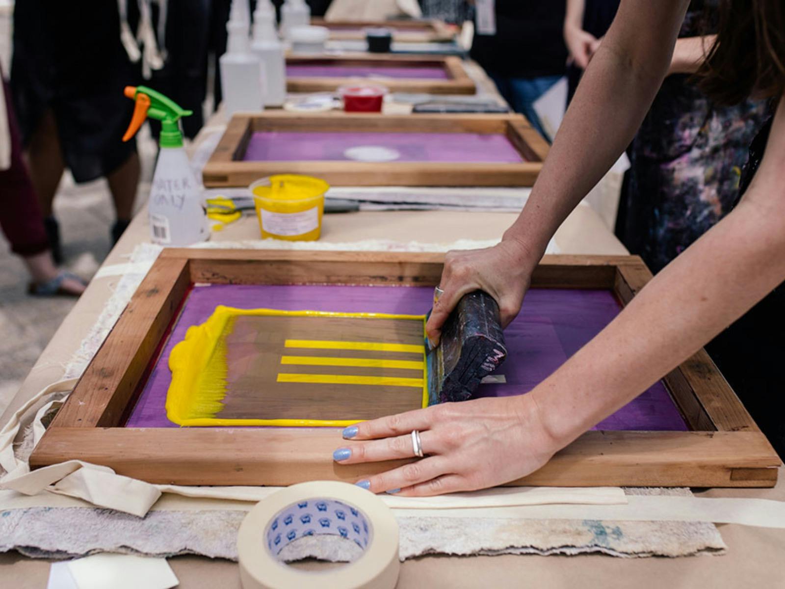 Hands rolling yellow ink onto a purple screen-printing frame with a squeegee at a workshop table.