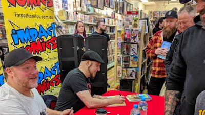 Two men sit at a table a line of people waiting to meet them