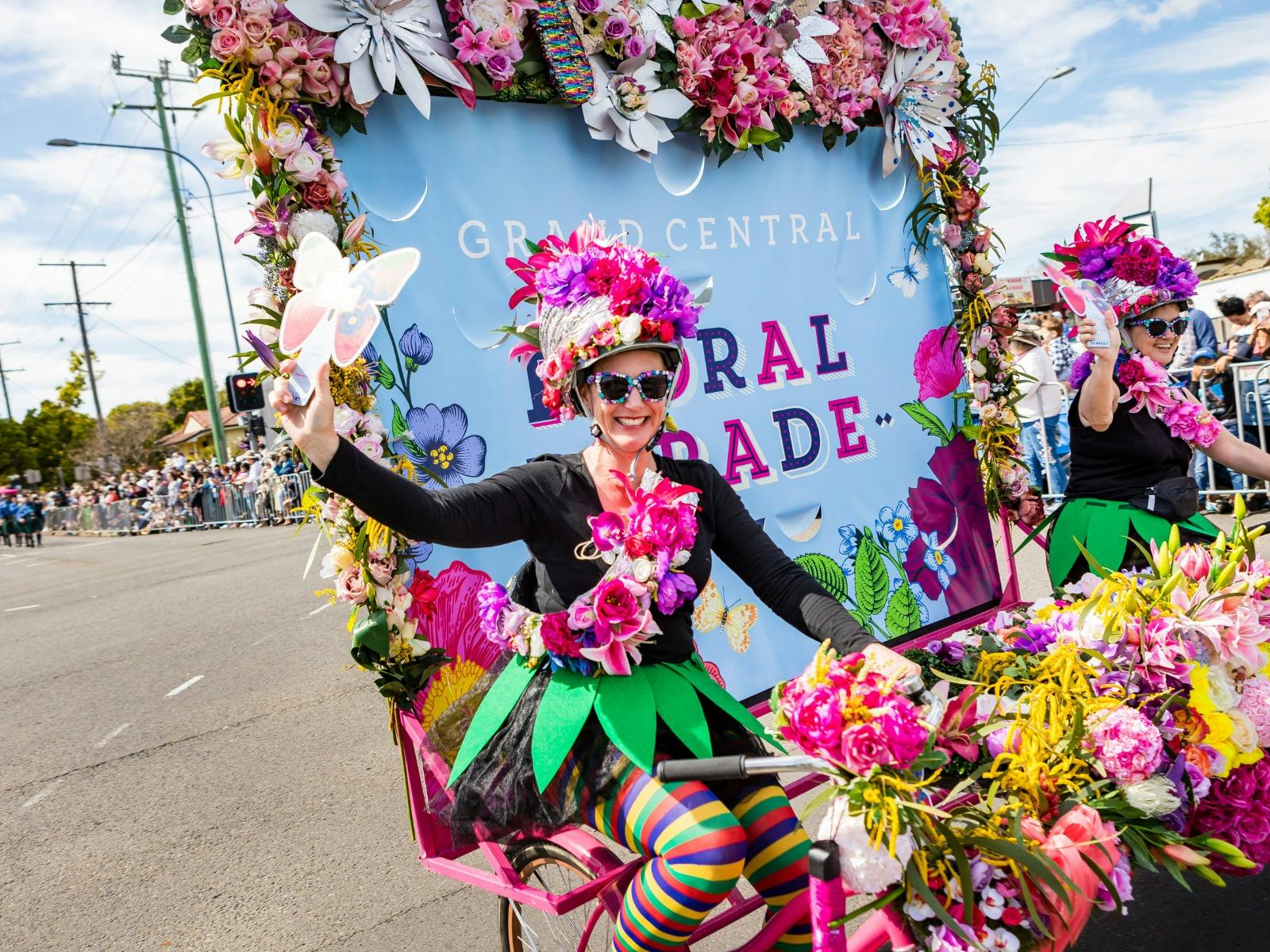 A brightly costumed woman rides a bike pulling a banner in a street parade.