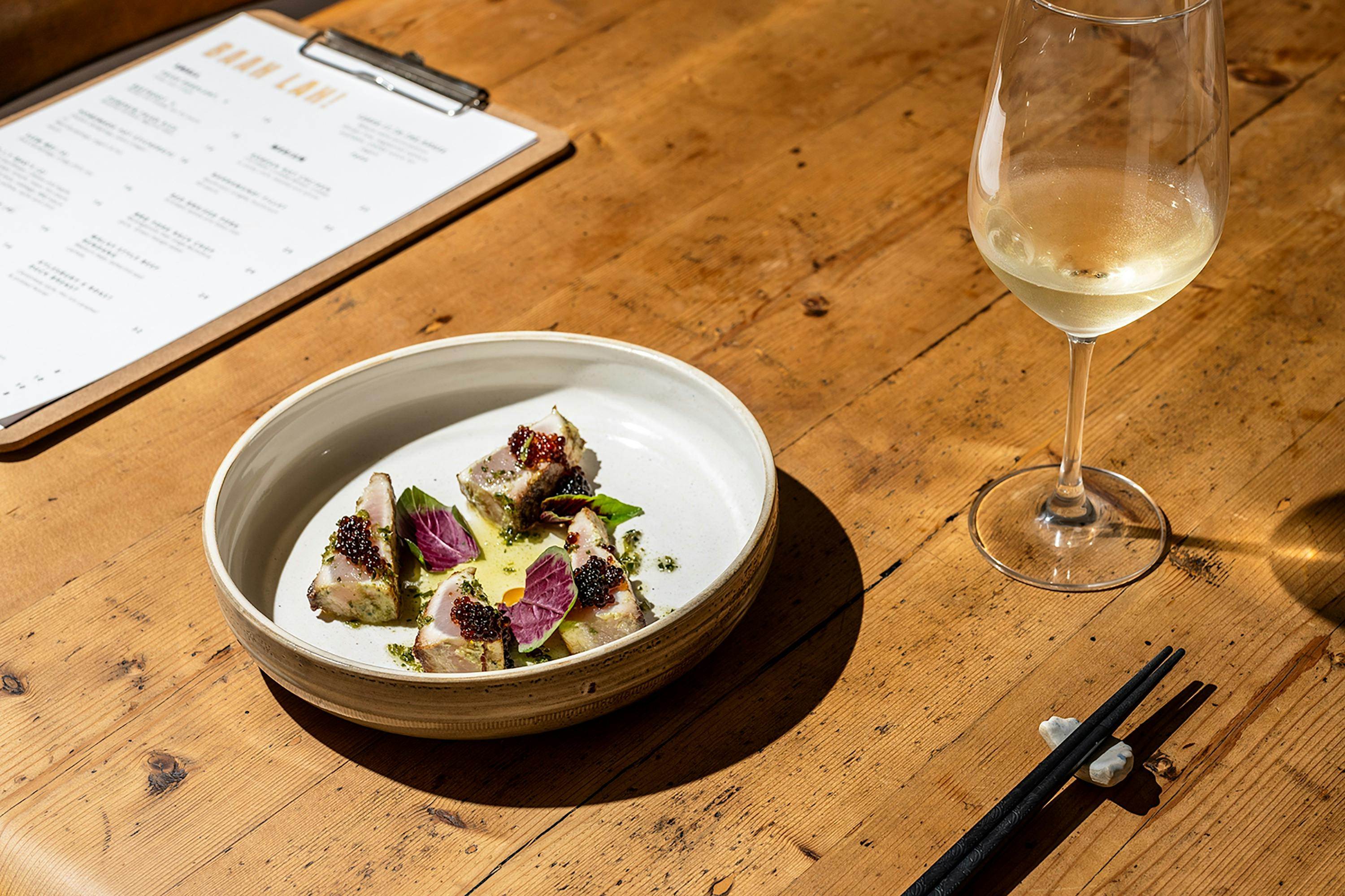 handmade ceramic bowl with seared kingfish pieces inside, a glass of white wine, a menu on  table