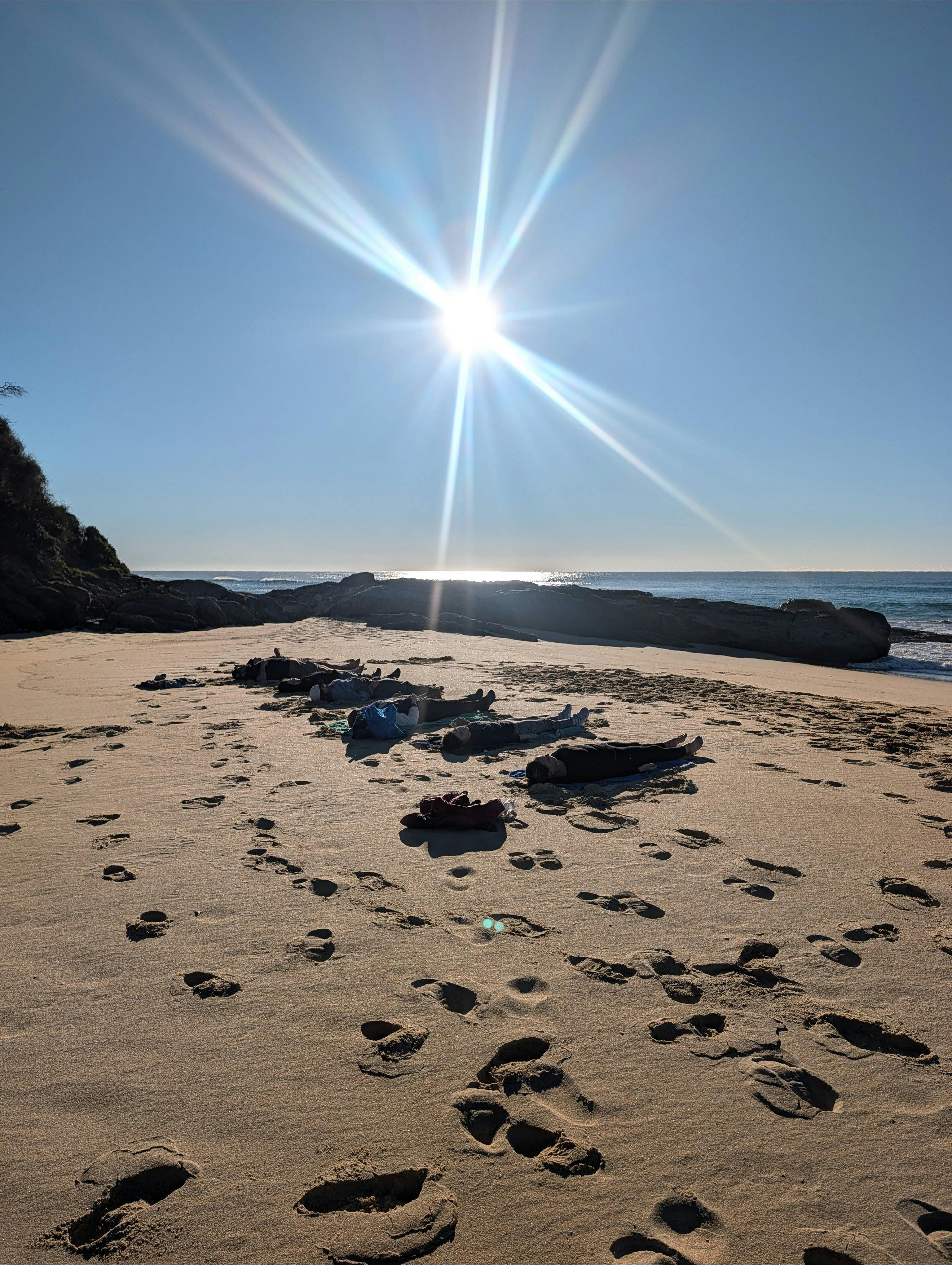 Students resting under a bright, star-like sunrise at barefoot beach yoga, Potato Point NSW.