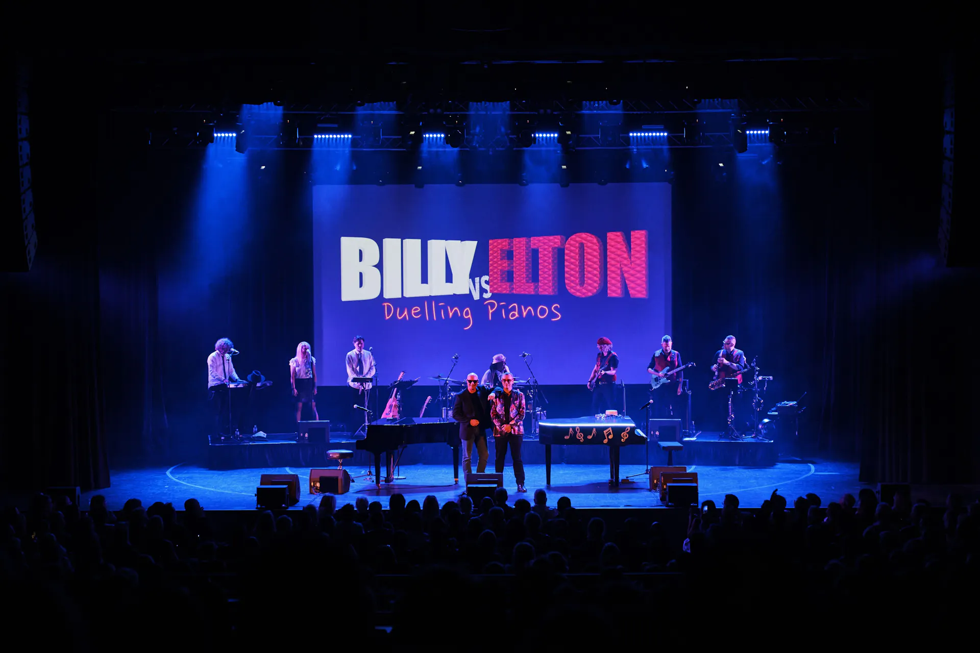 A stage with blue lights streaming from above. A band is on stage with 'Billy Vs Elton' projected