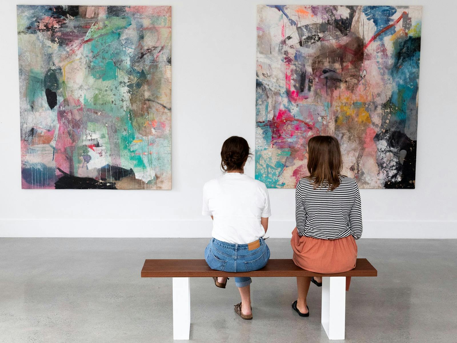 Two people are sitting on a bench in the gallery looking large colourful abstract paintings