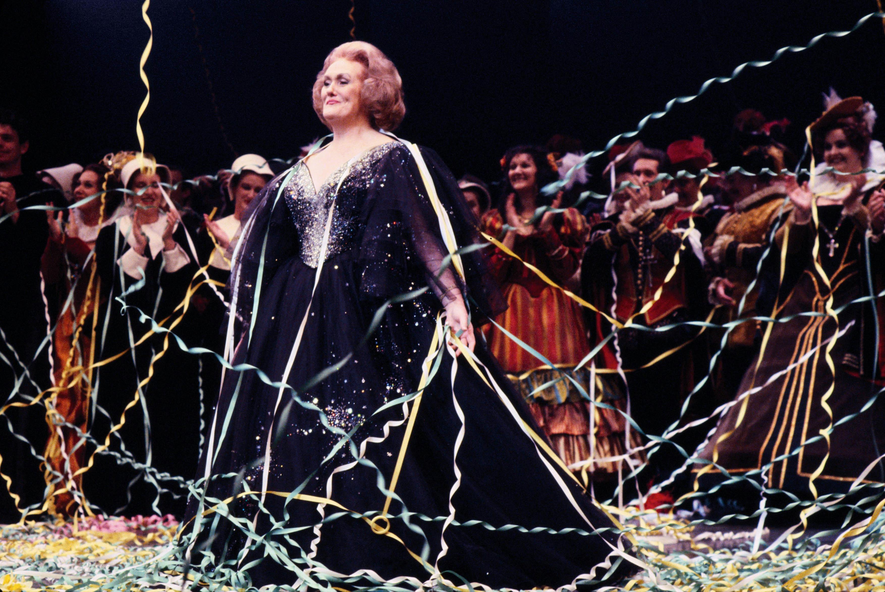 Dame Joan Sutherland stands at curtain call surrounded by streamers. A chorus behind applauds her.