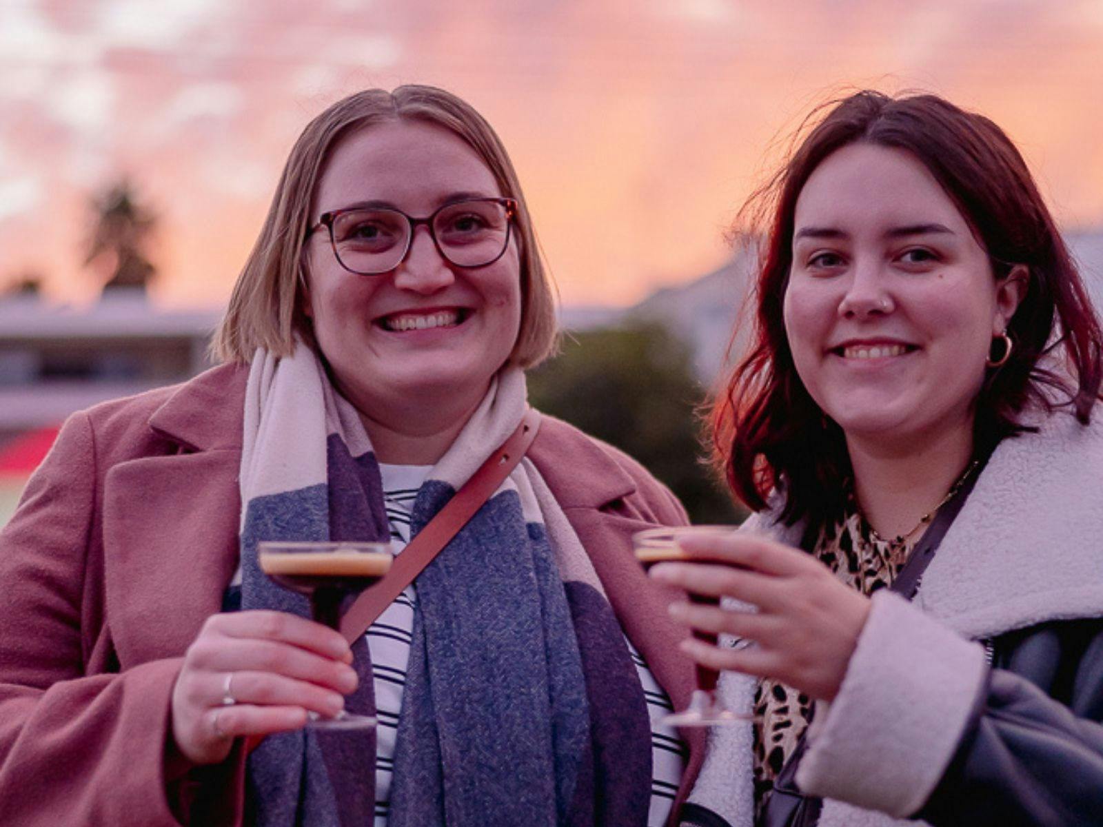 Two ladies smiling, each holding an expresso martini cocktail with sunset in background
