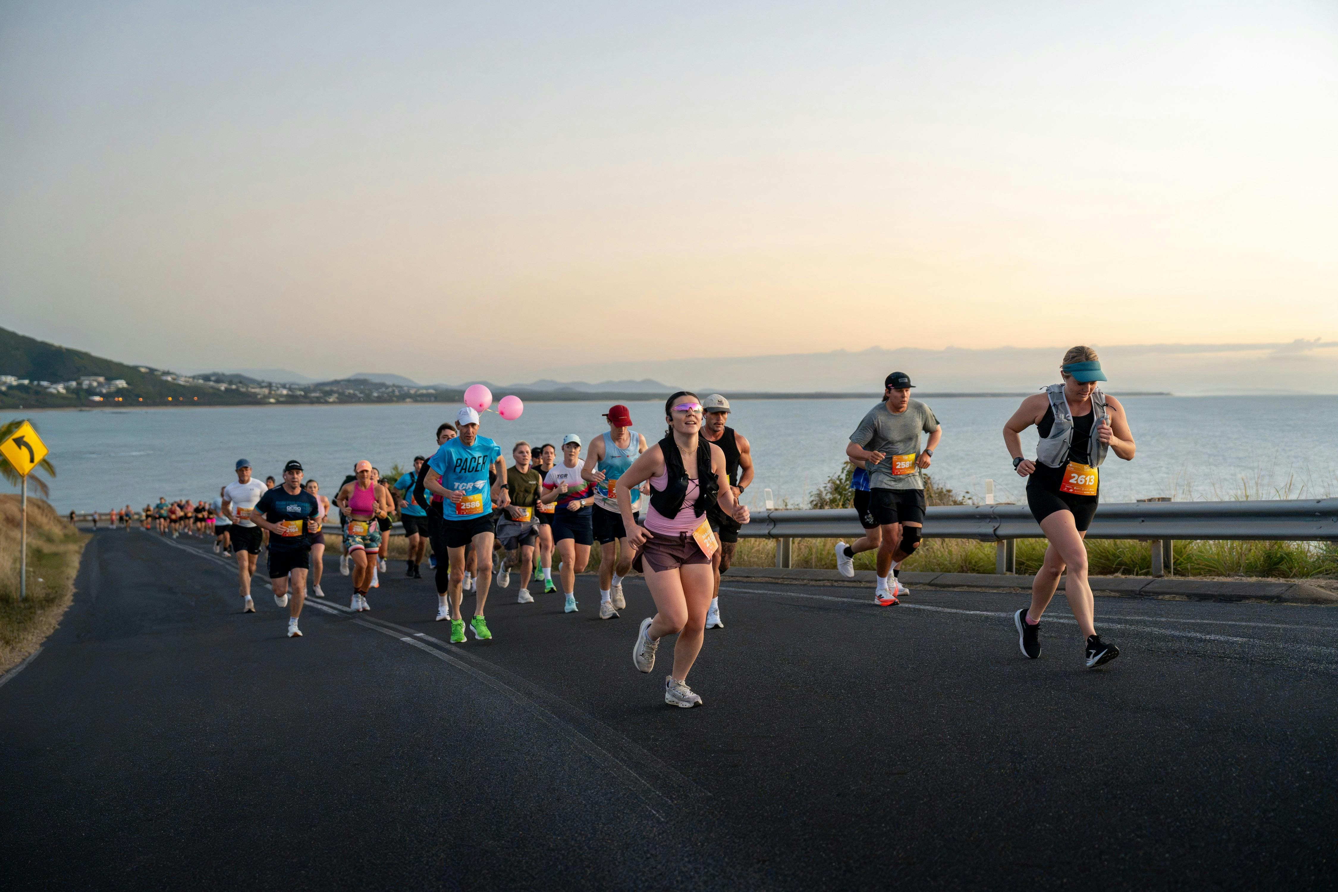 A large group of runners travels along a coastal road at sunrise, with the ocean and Yeppoon coastli