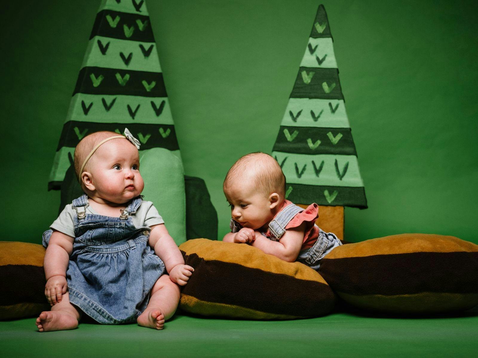 Two babies sit on some log cushions with soft cartoon trees behind them