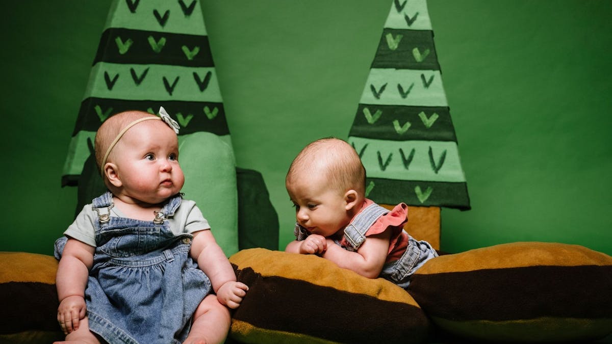 Two babies sit on some log cushions with soft cartoon trees behind them