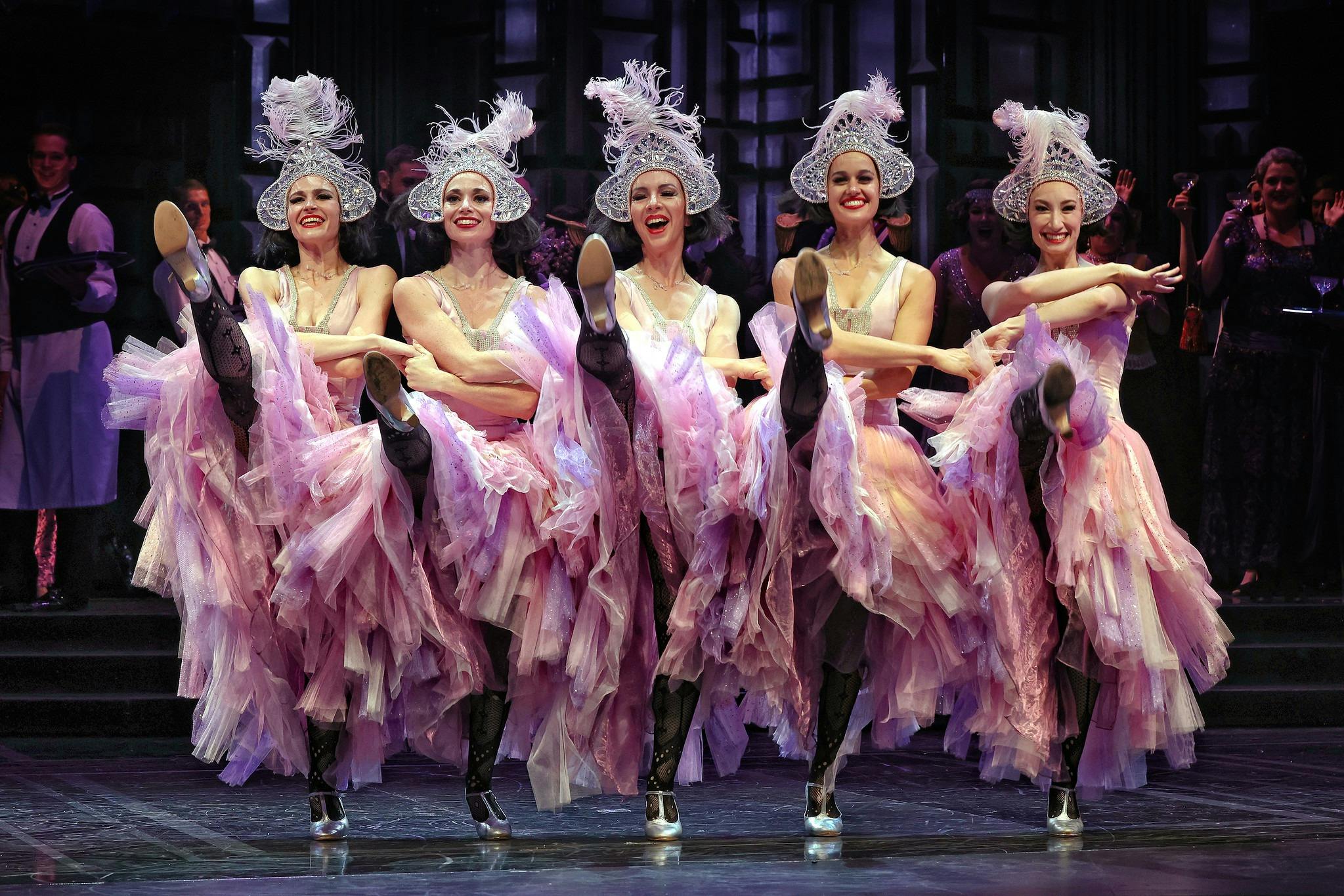 Five dancers in pink feathered costumes kick in unison onstage, smiling in chorus line.