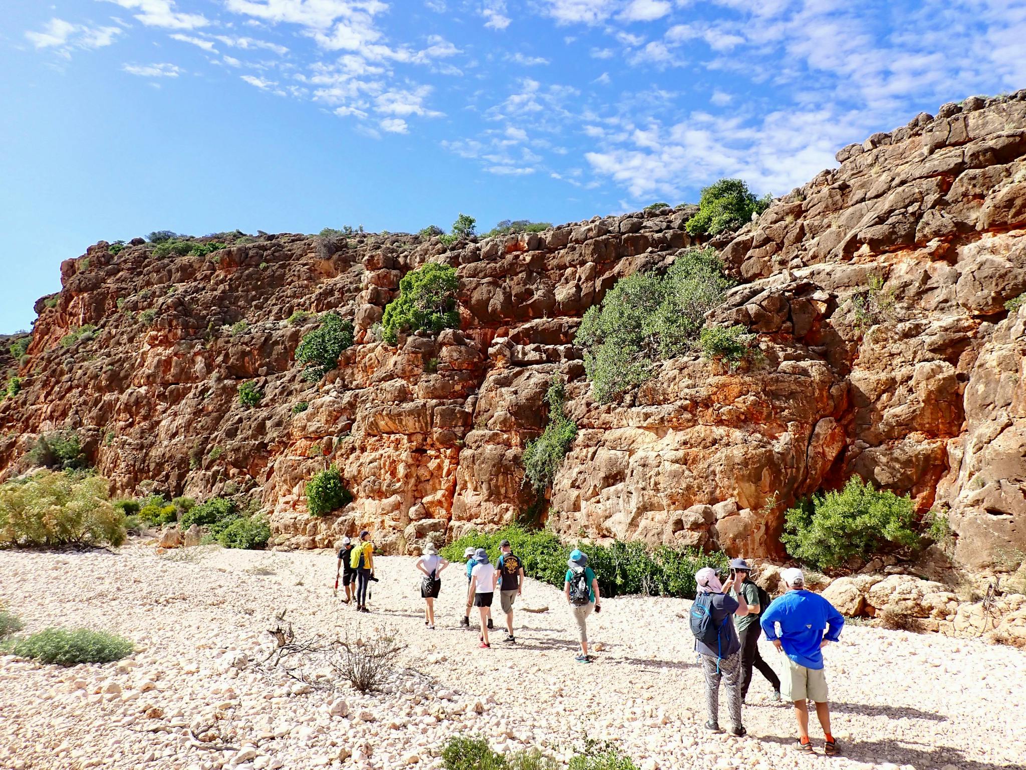 A group of people standing at the base of a sheer red rock cliff admiring the view
