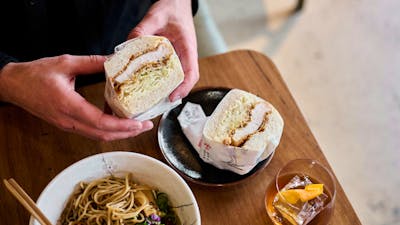 Hands holding a fluffy katsu sandwich with a bowl or ramen and a drink nearby.