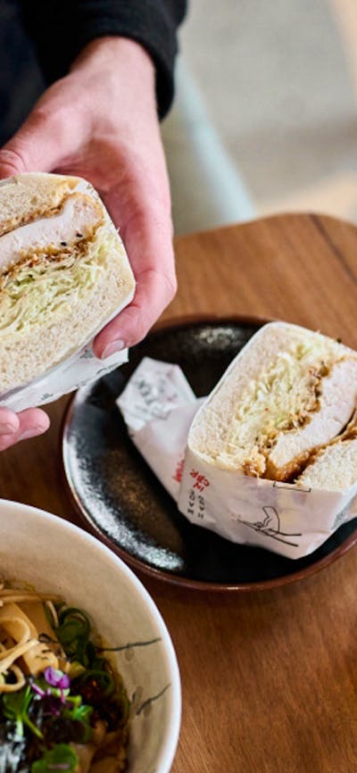 Hands holding a fluffy katsu sandwich with a bowl or ramen and a drink nearby.