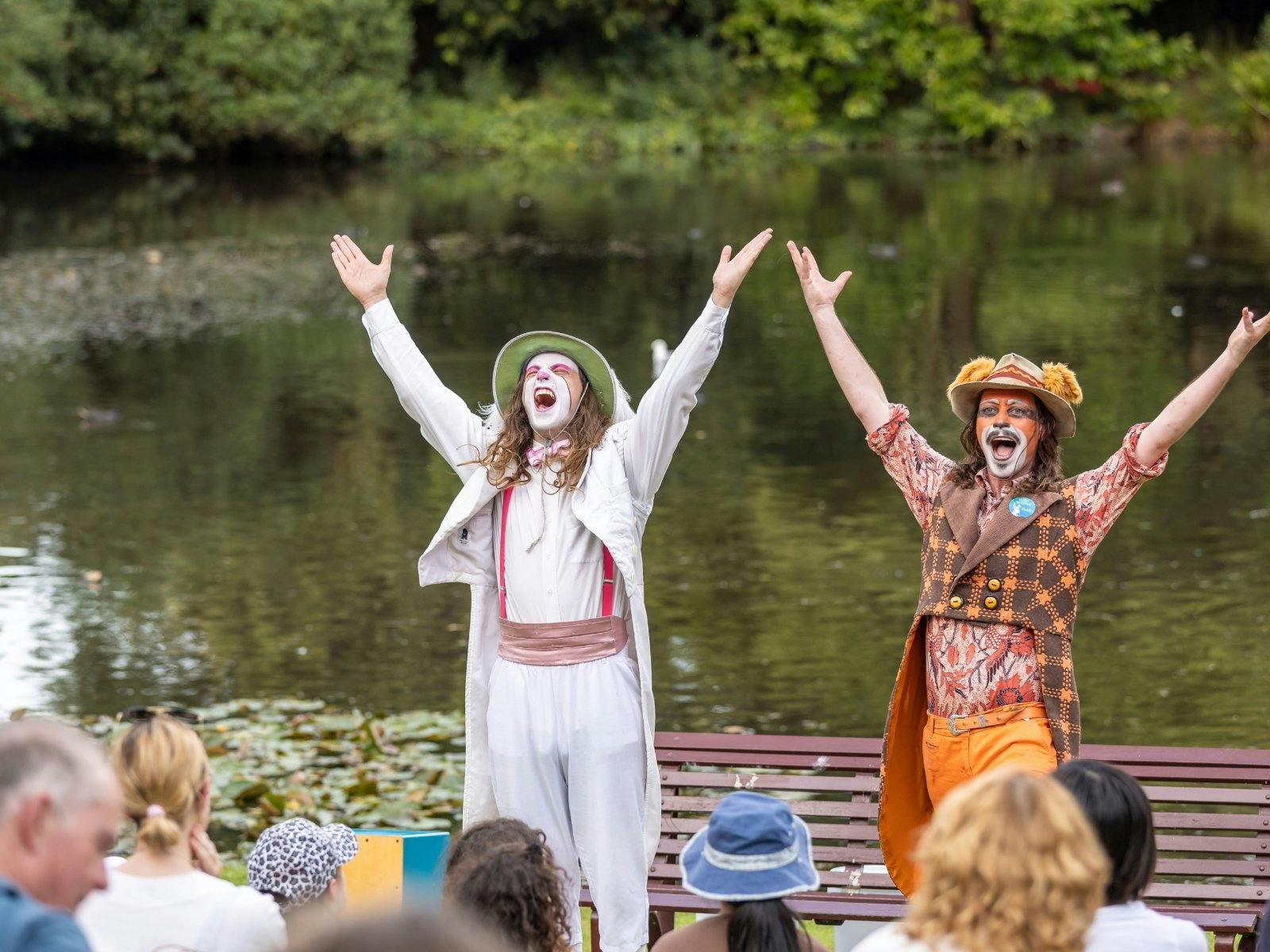 Two performers dressed as animals with arms up by a lake, performing in front of an audience.