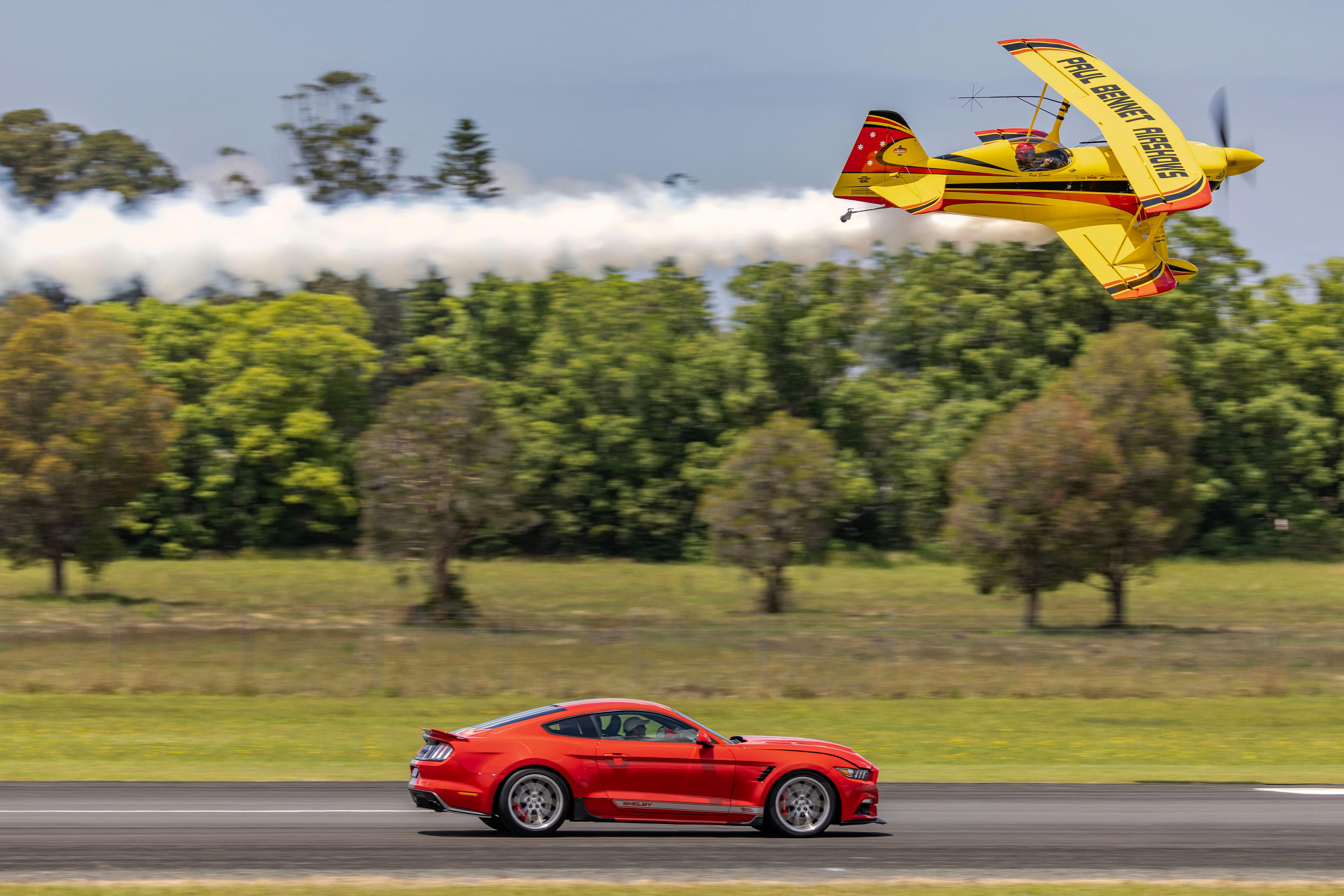 The Wolf Pitts Pro biplane makes a low-level pass above a red Shelby Mustang on the runway