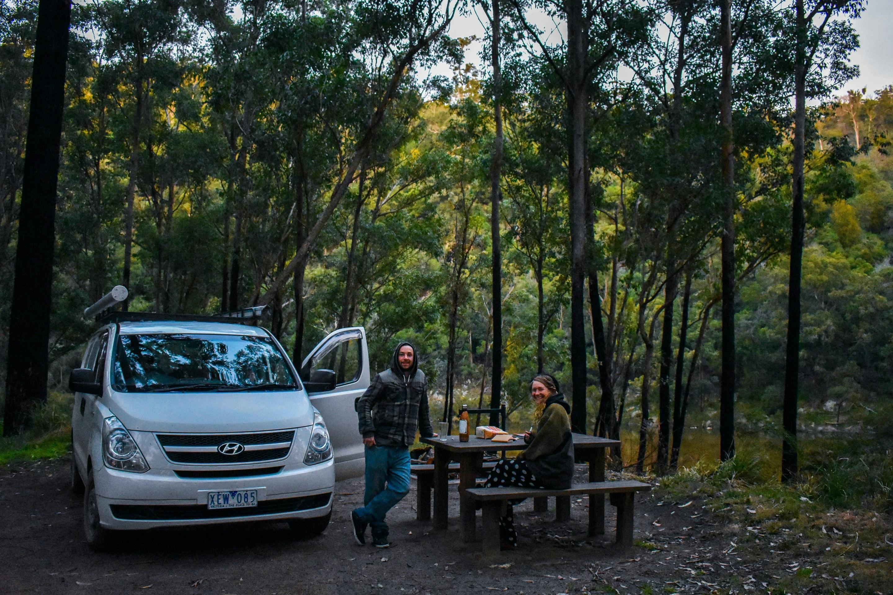 Two people with a car at a picnic table surrounded by forest