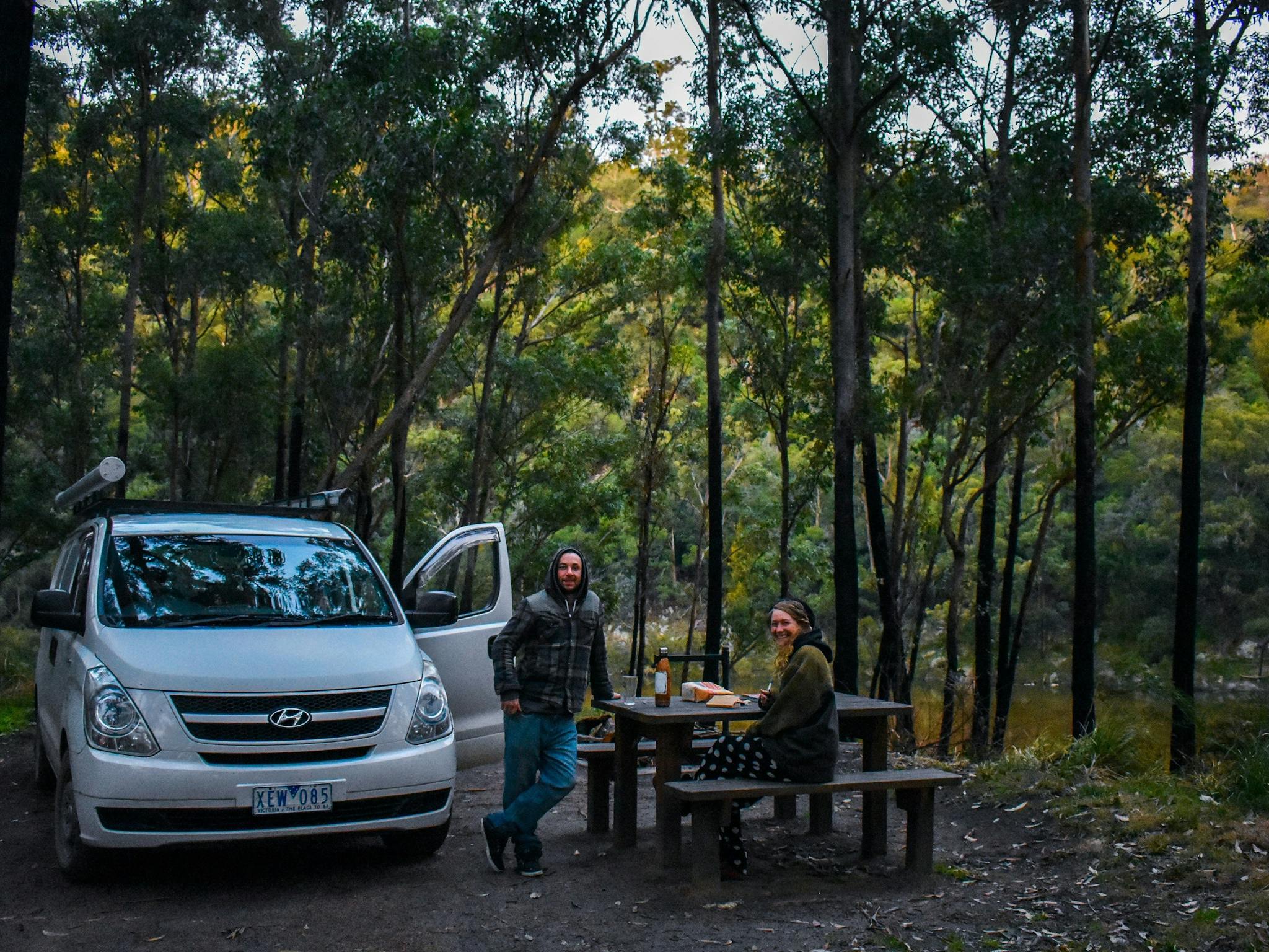 Two people with a car at a picnic table surrounded by forest