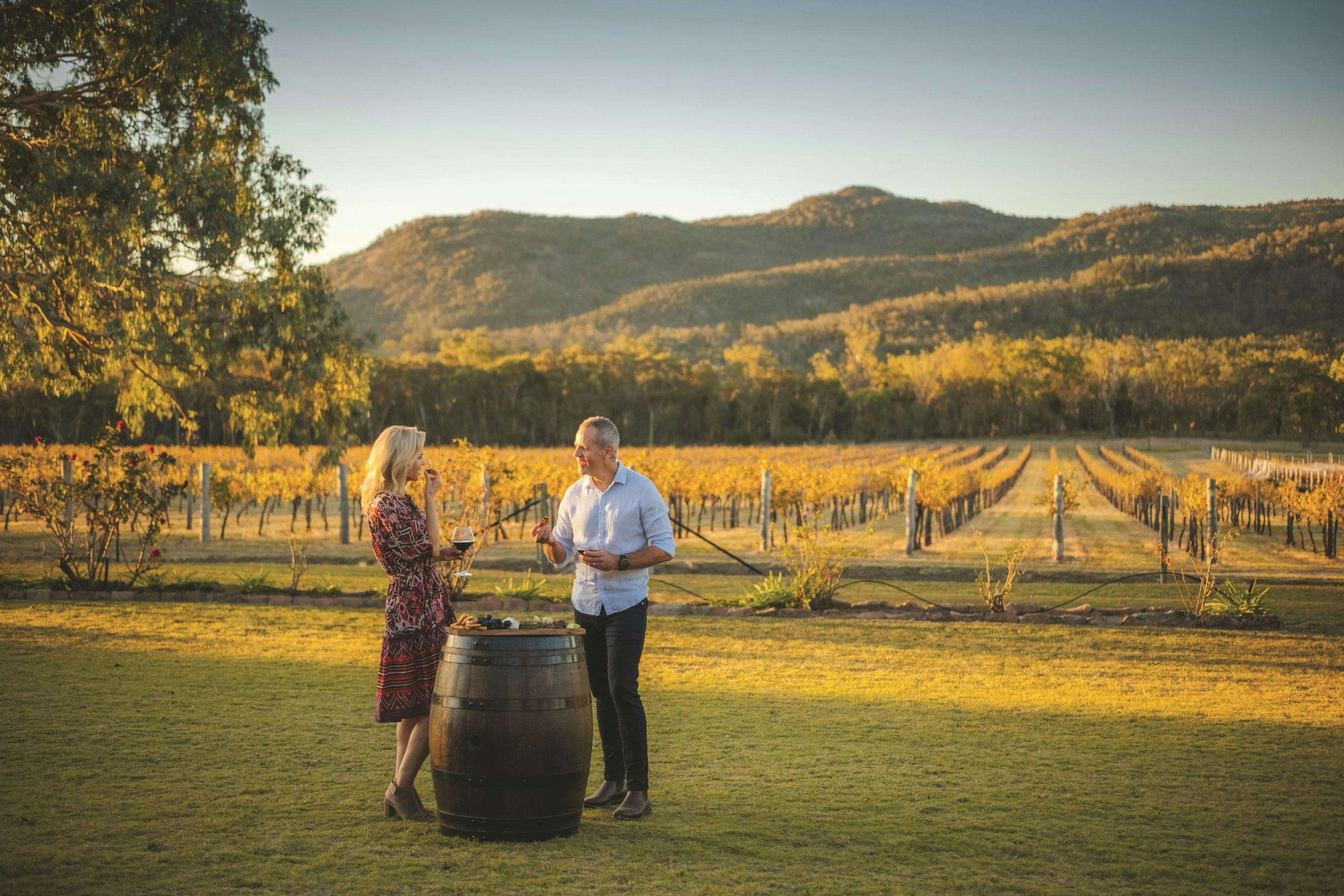 Man and woman standing at a wine barrel with red wine and cheese platter vineyard in background