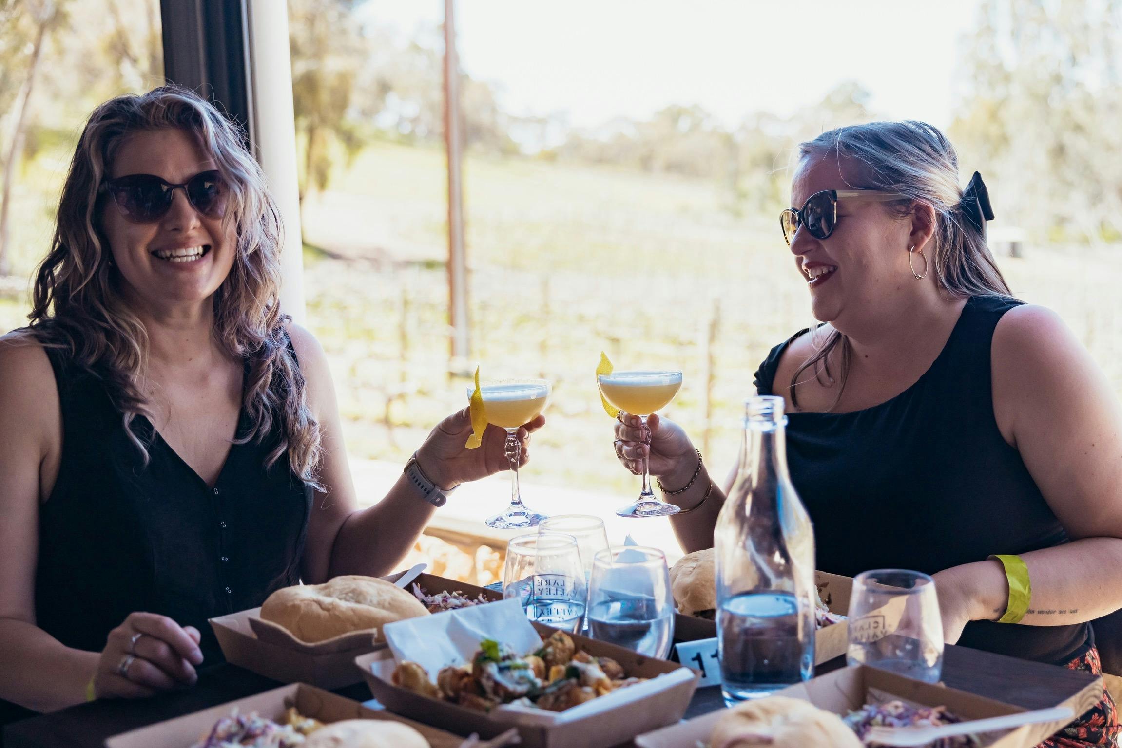 2 ladies holding cocktail glasses and smiling with vines in the background