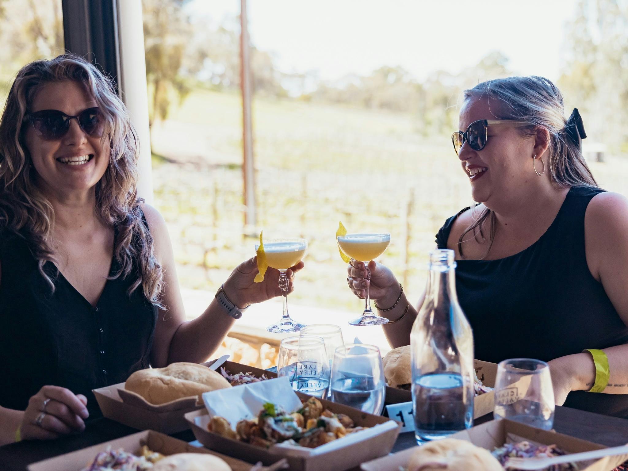 2 ladies holding cocktail glasses and smiling with vines in the background