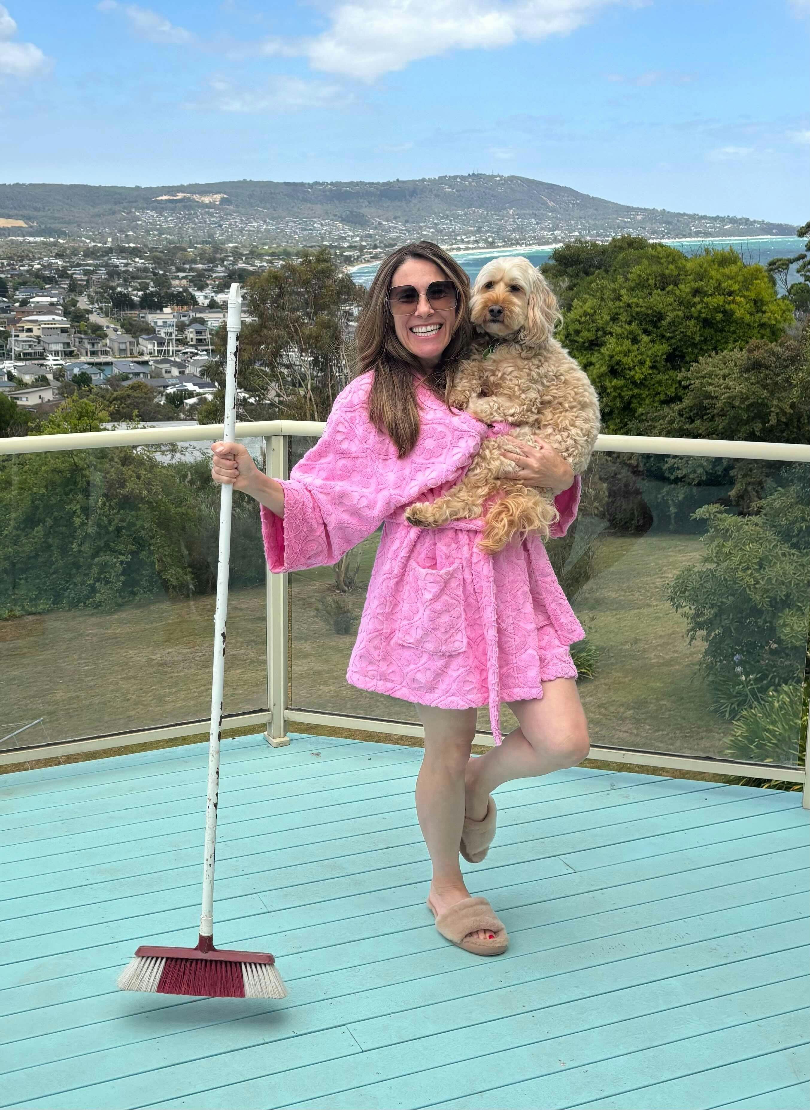 Marilyn is on a blue balcony, wearing a pink bathrobe and sunglasses, holding a broom and small dog
