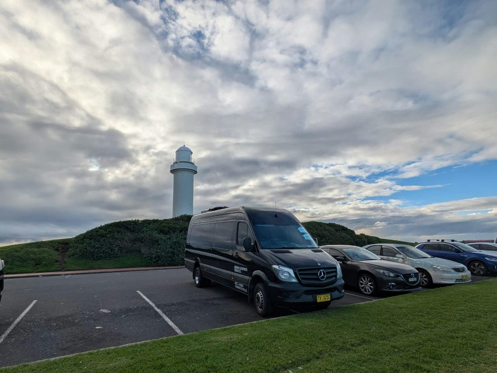 Black Mercedes Sprinter minibus parked below the white Wollongong lighthouse on a cloudy day
