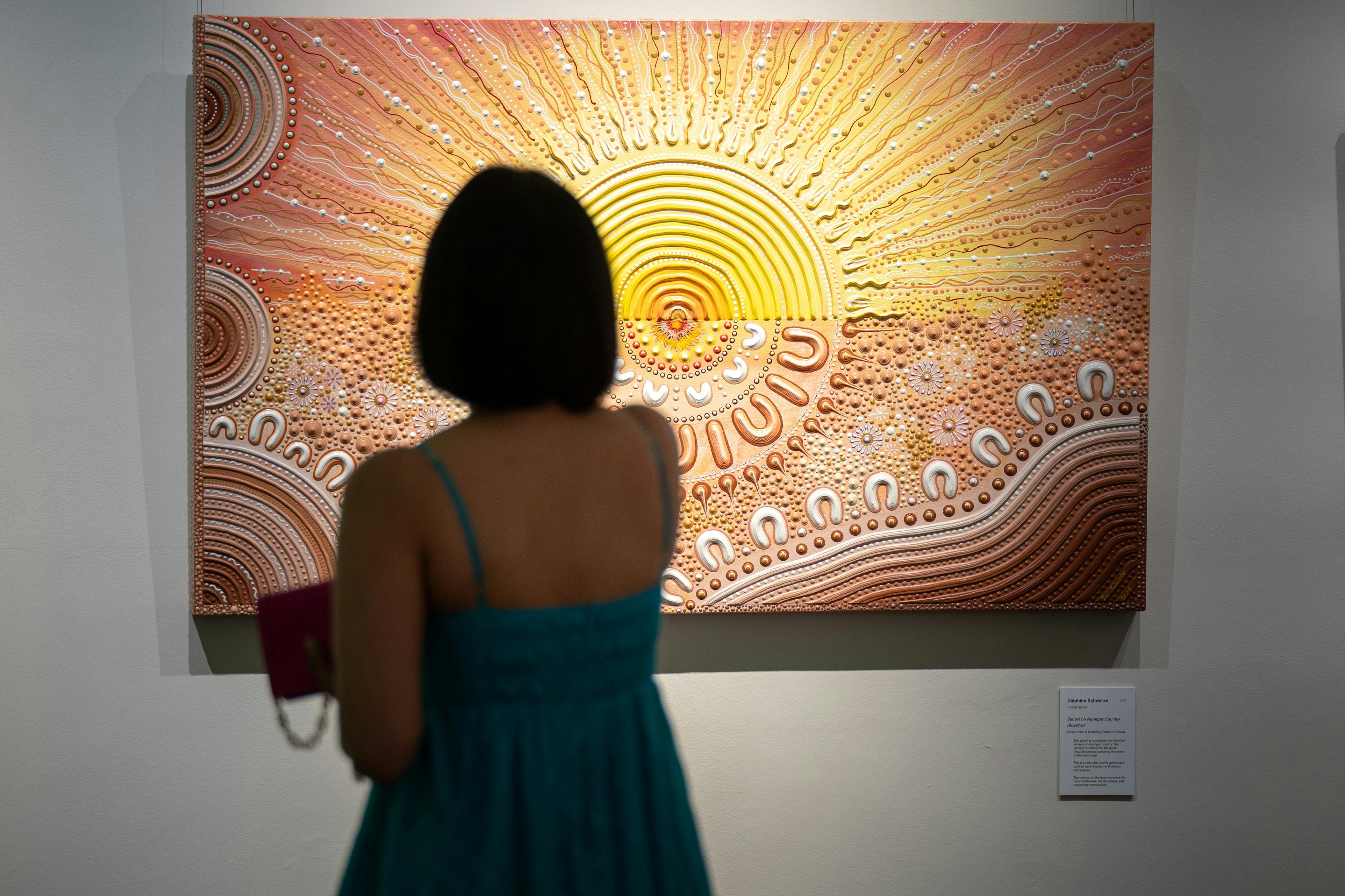 A woman in a teal dress views a vibrant, textured painting of a radiant sun at an art gallery.