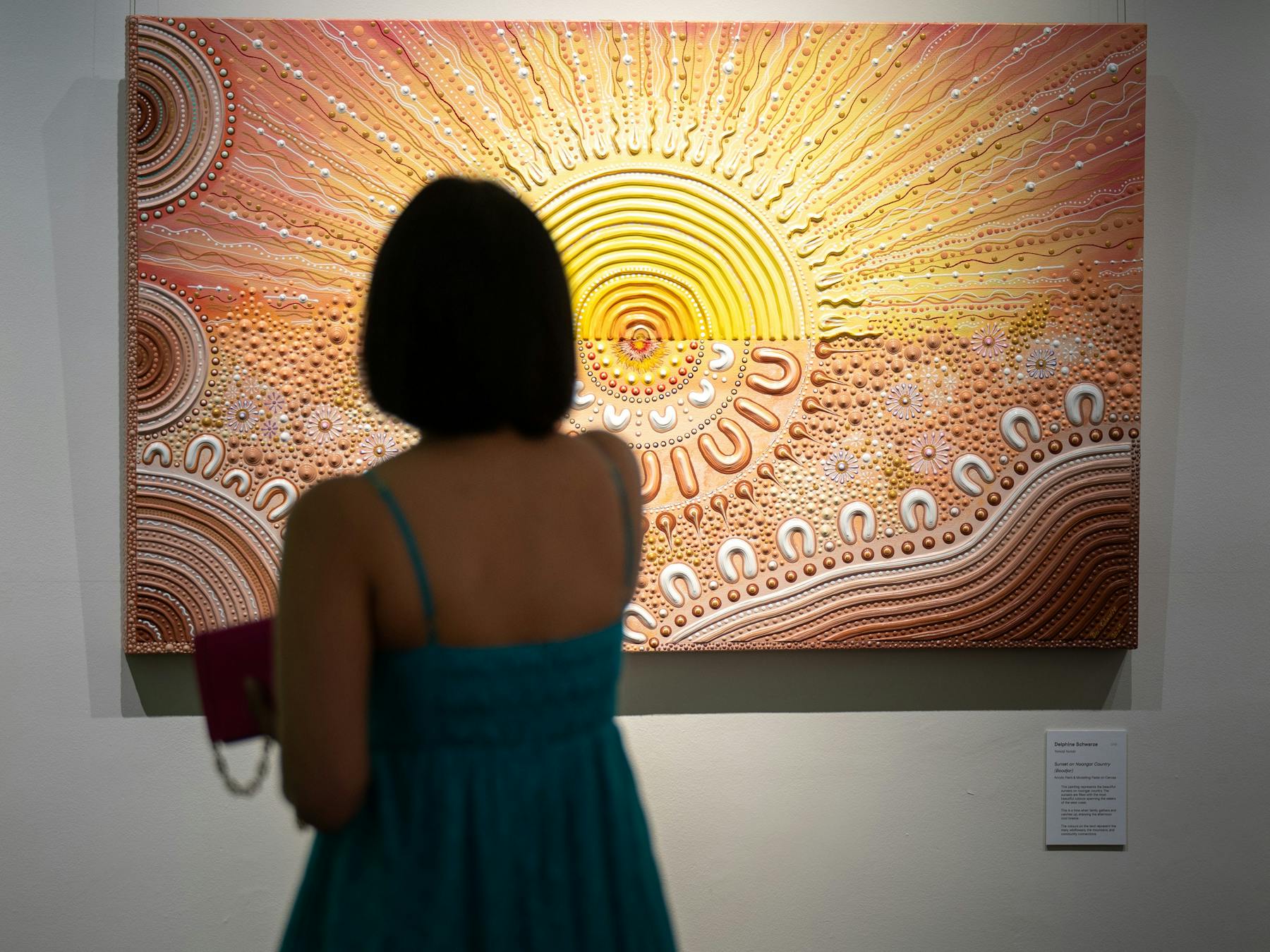 A woman in a teal dress views a vibrant, textured painting of a radiant sun at an art gallery.