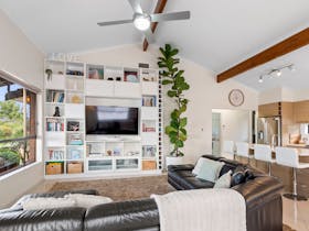 Spacious living area with sofa, TV shelving, ceiling fan, kitchen island seating, and large windows.