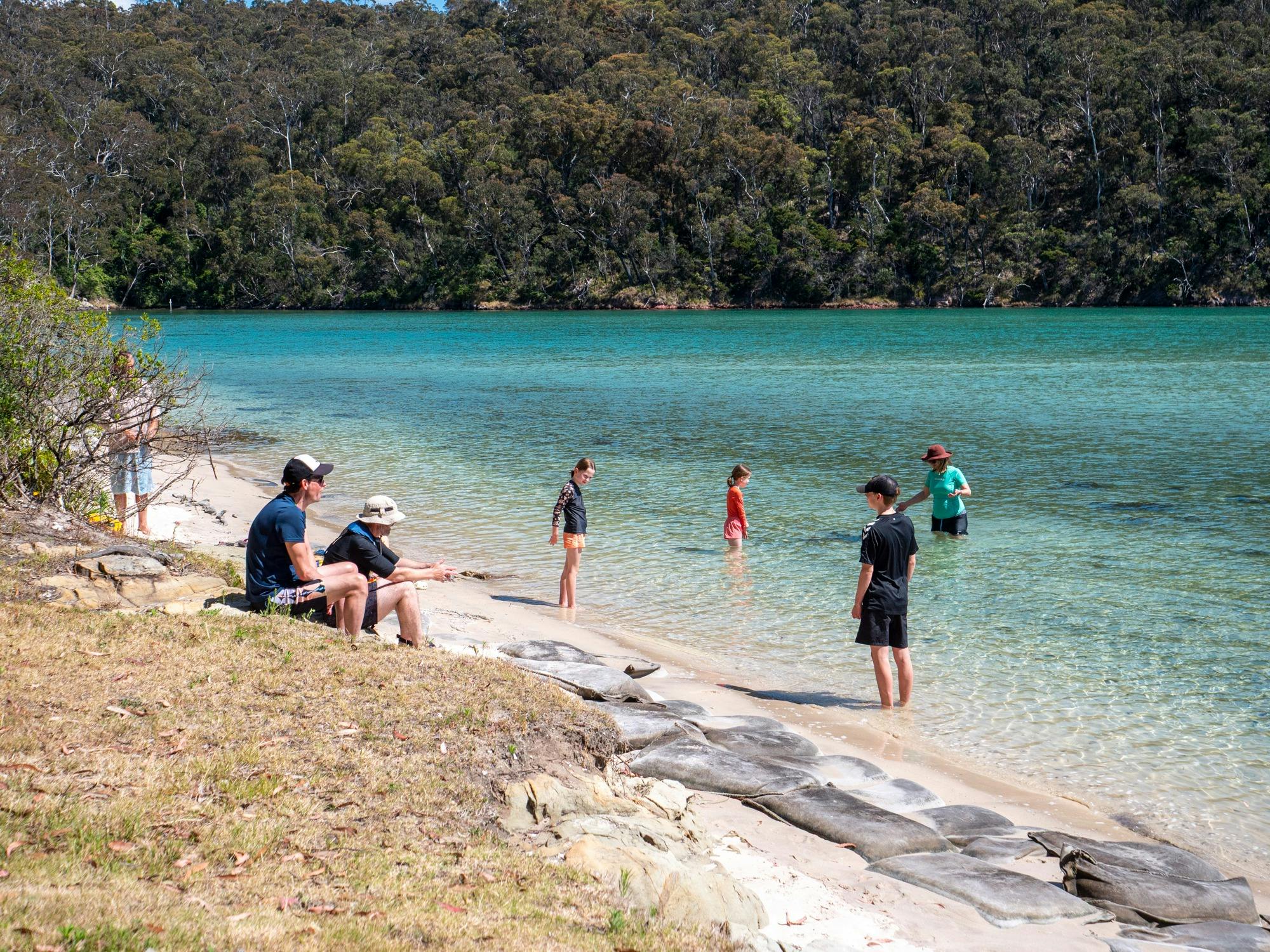 Children swimming in turquoise Pambula River water with a sandy beach in the foreground.