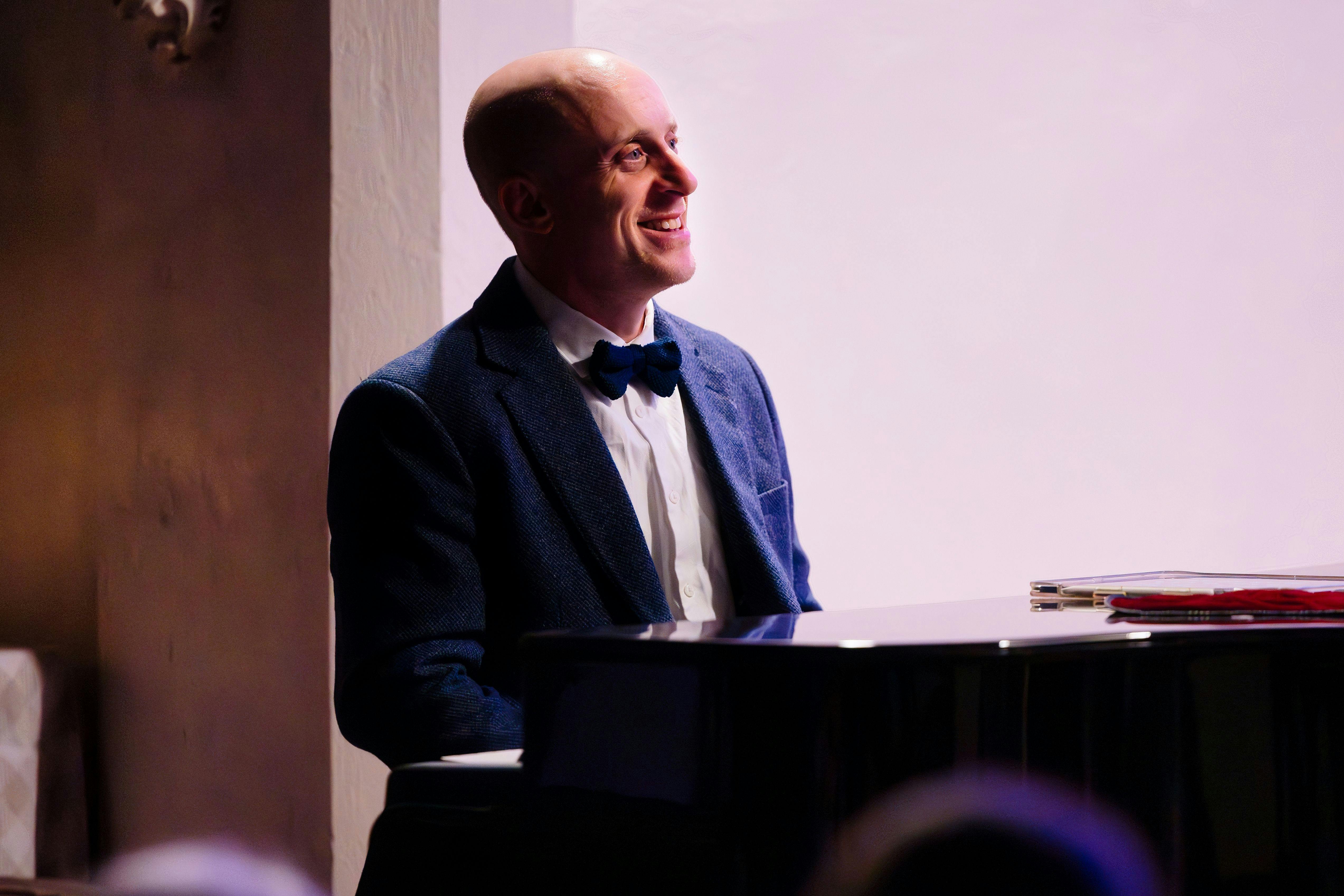 a man in a dinner jacket sits at a baby grand piano. The background lighting is pale pink