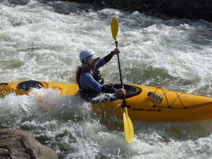 A girl is paddling in whitewater on the snowy, wearing a helmet and fully covered against sun burn