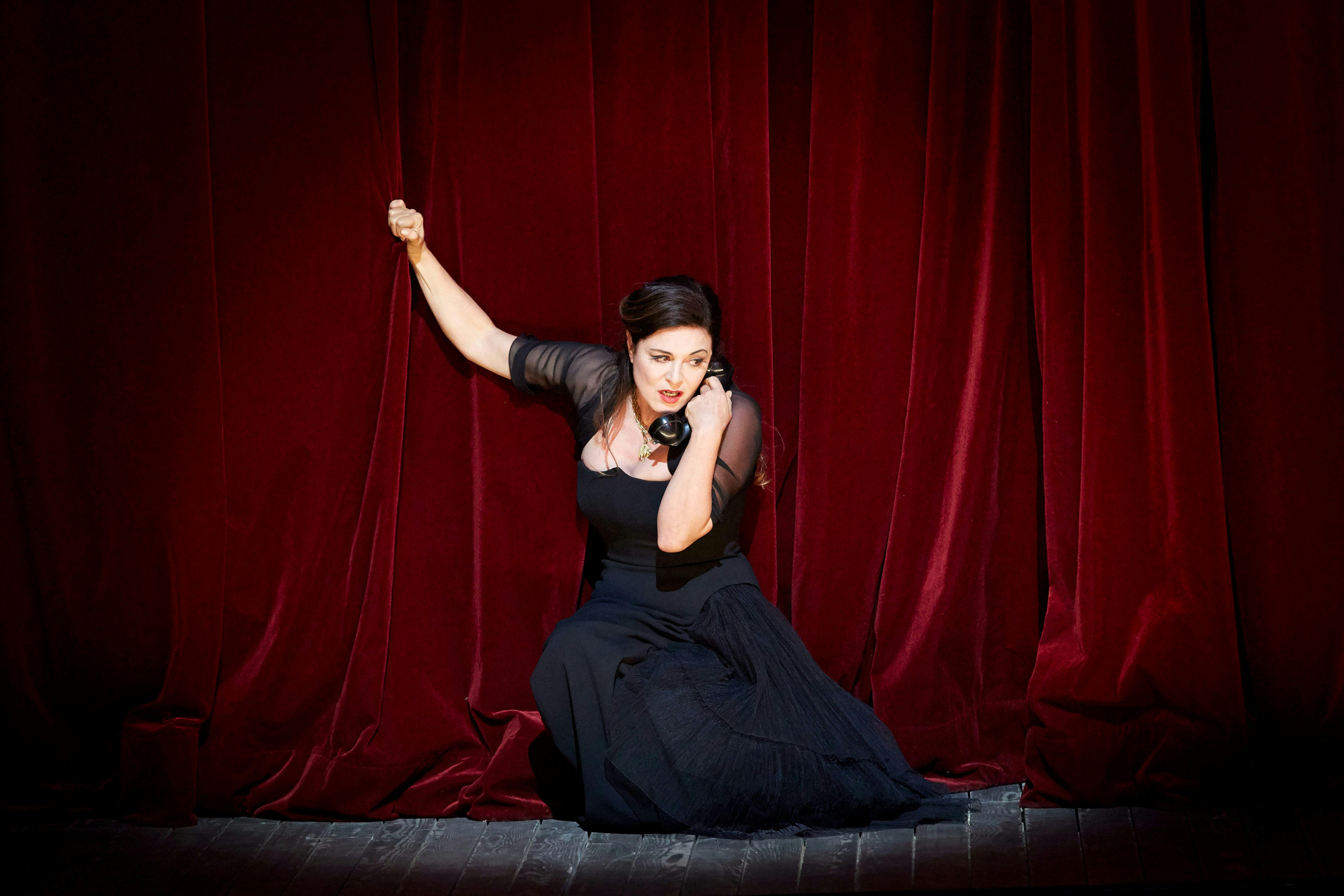 A woman in a black gown on the telephone grips a red curtain backdrop as she falls to her knees