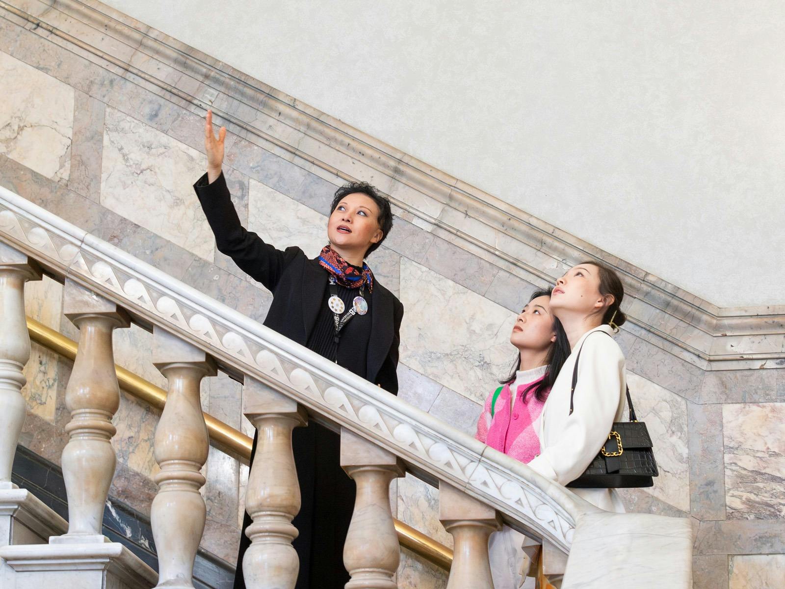 Two people are led up a grand marble staircase by a tour guide.