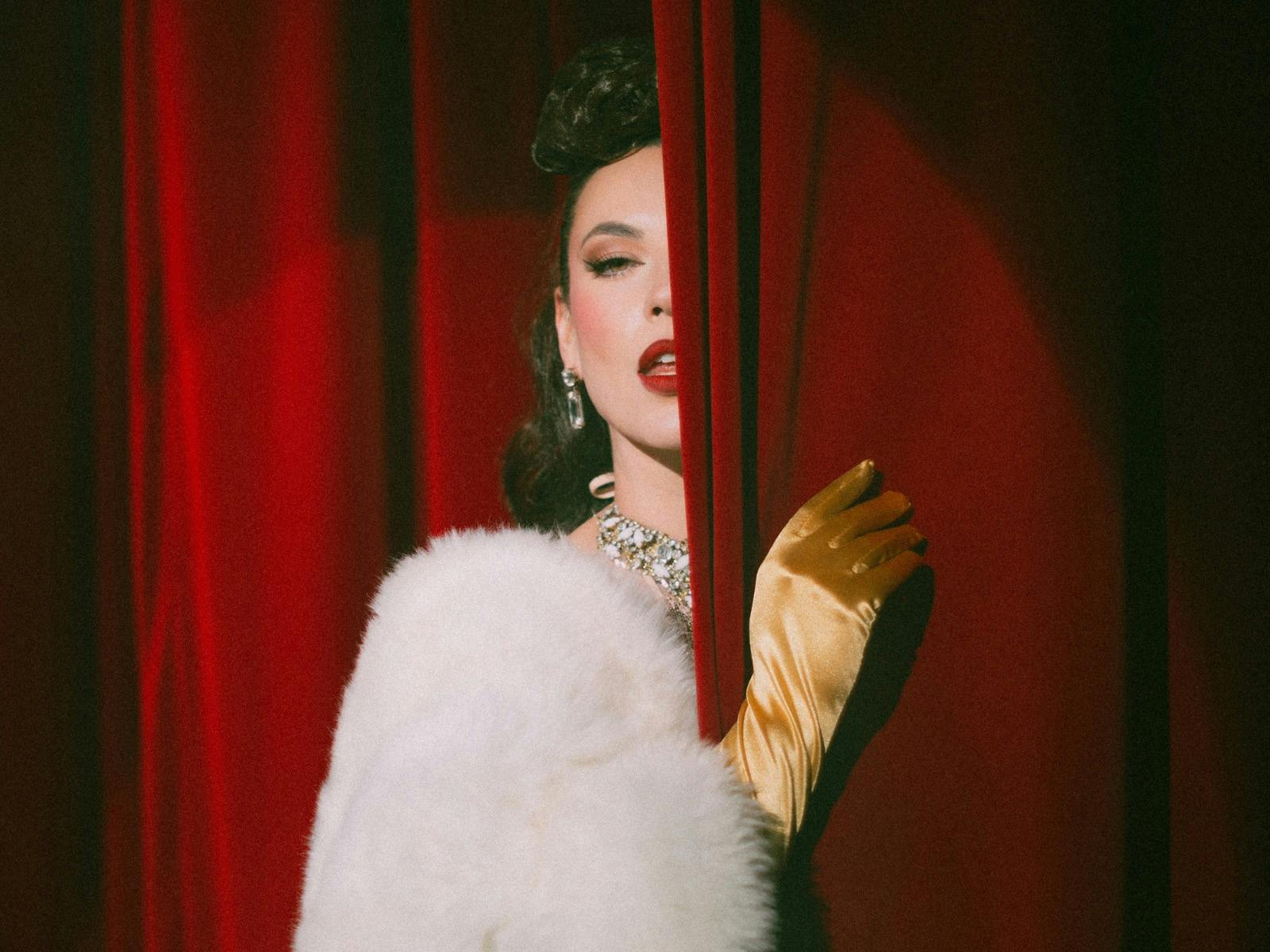 A seductive brunette burlesque performer peaks behind a velvet red curtain