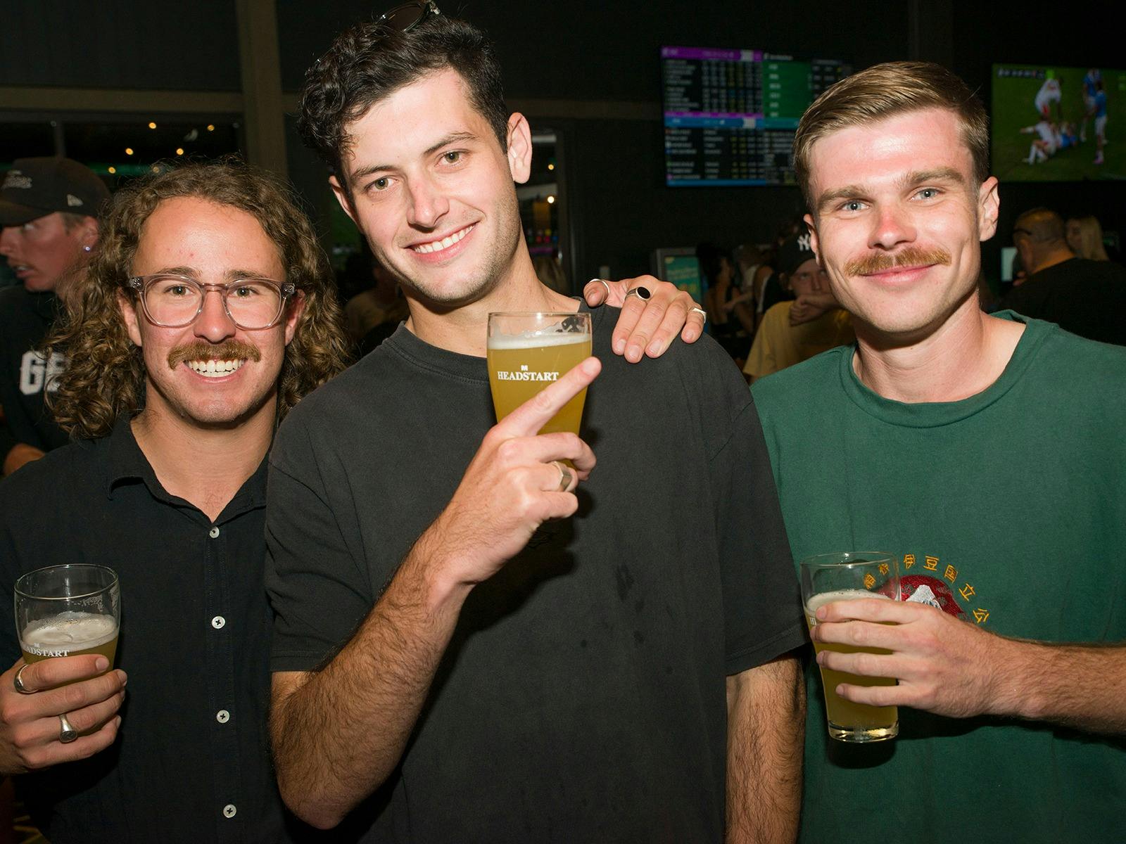 Three men standing next to one another holding schooners of beer and smiling at the camera