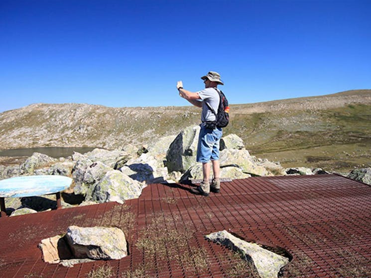 A man takes a photo at Cootapatamba lookout, with Mount Kosciuszko in the background, Kosciuszko