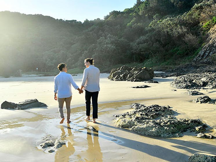 A couple walking hand-in-hand along the sand at Tallow Beach in Cape Byron State Conservation Area.