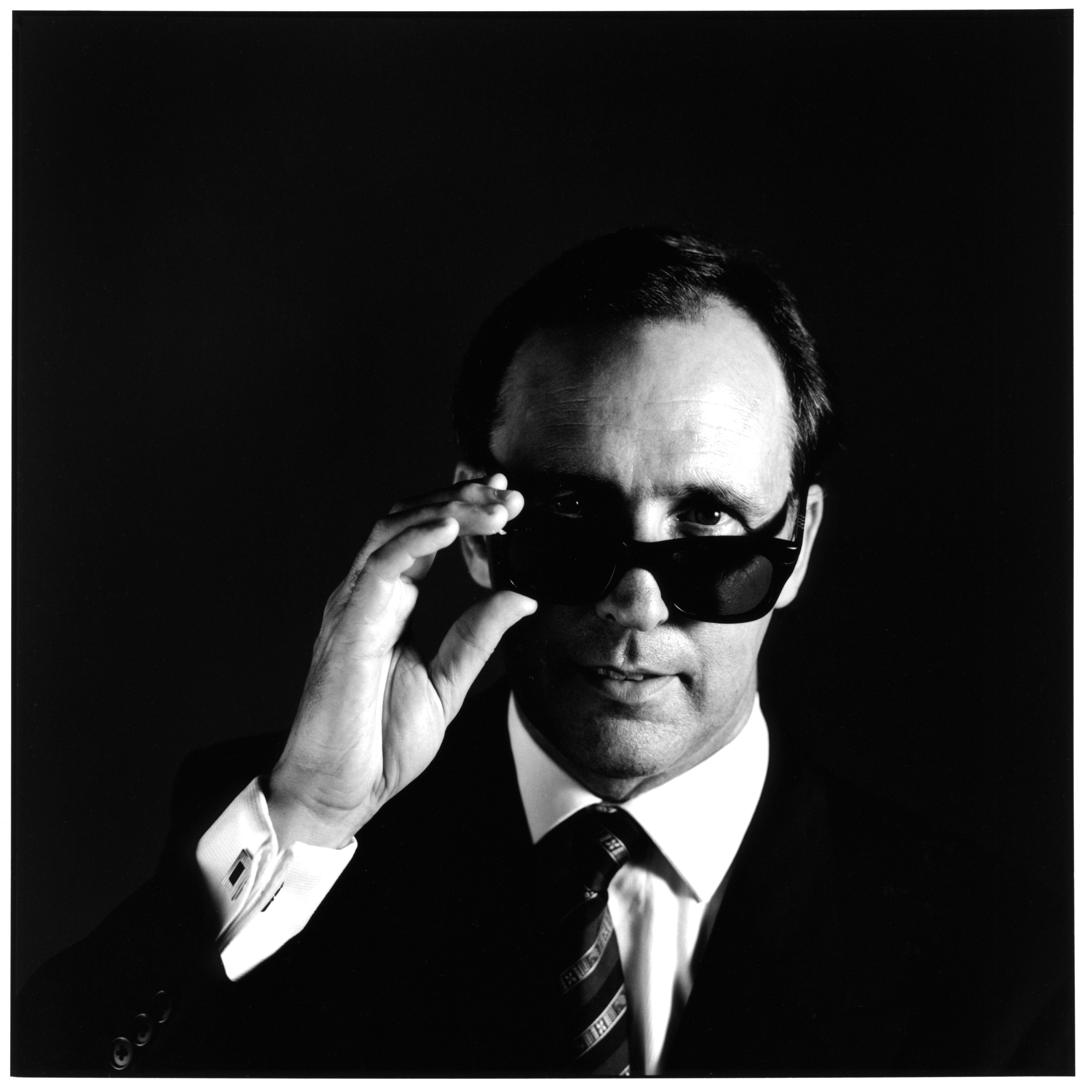 A black and white photo of a man holding his hand up to his sunglasses