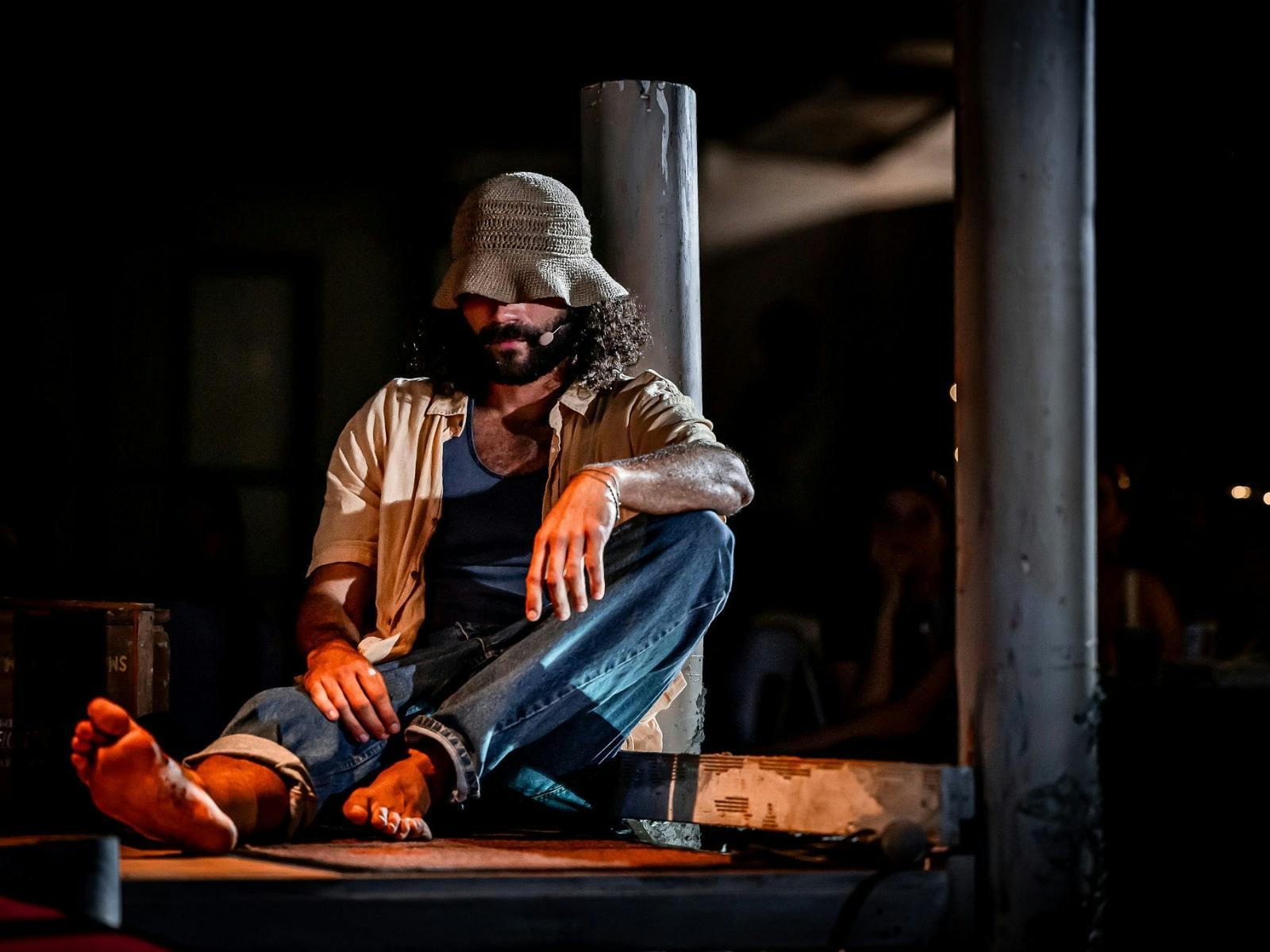 Actor sits barefoot on stage floor, bucket hat low, wearing headset mic, leaning against a pole.